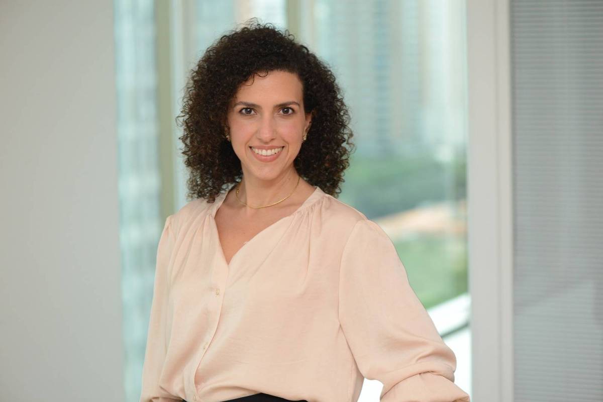 A imagem mostra uma mulher com cabelo cacheado e escuro, usando uma blusa clara. Ela está sorrindo e posando em um ambiente interno com janelas grandes ao fundo, que mostram uma vista urbana. A iluminação é suave e natural, destacando seu rosto e a blusa que ela usa.
