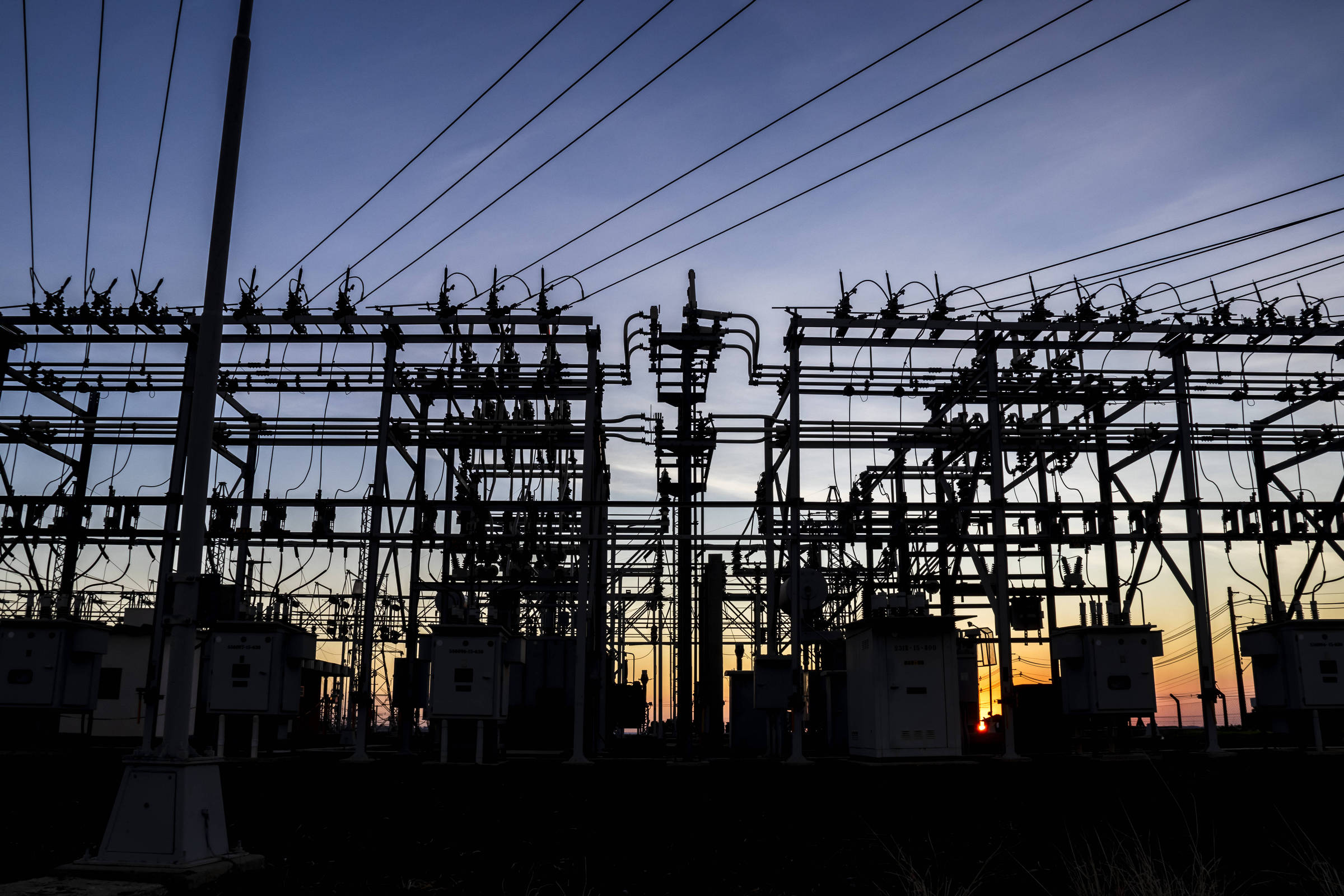 A imagem mostra uma subestação elétrica em silhueta durante o entardecer. Estruturas metálicas, transformadores e fios de alta tensão são visíveis, com um céu colorido ao fundo, apresentando tons de azul e laranja.