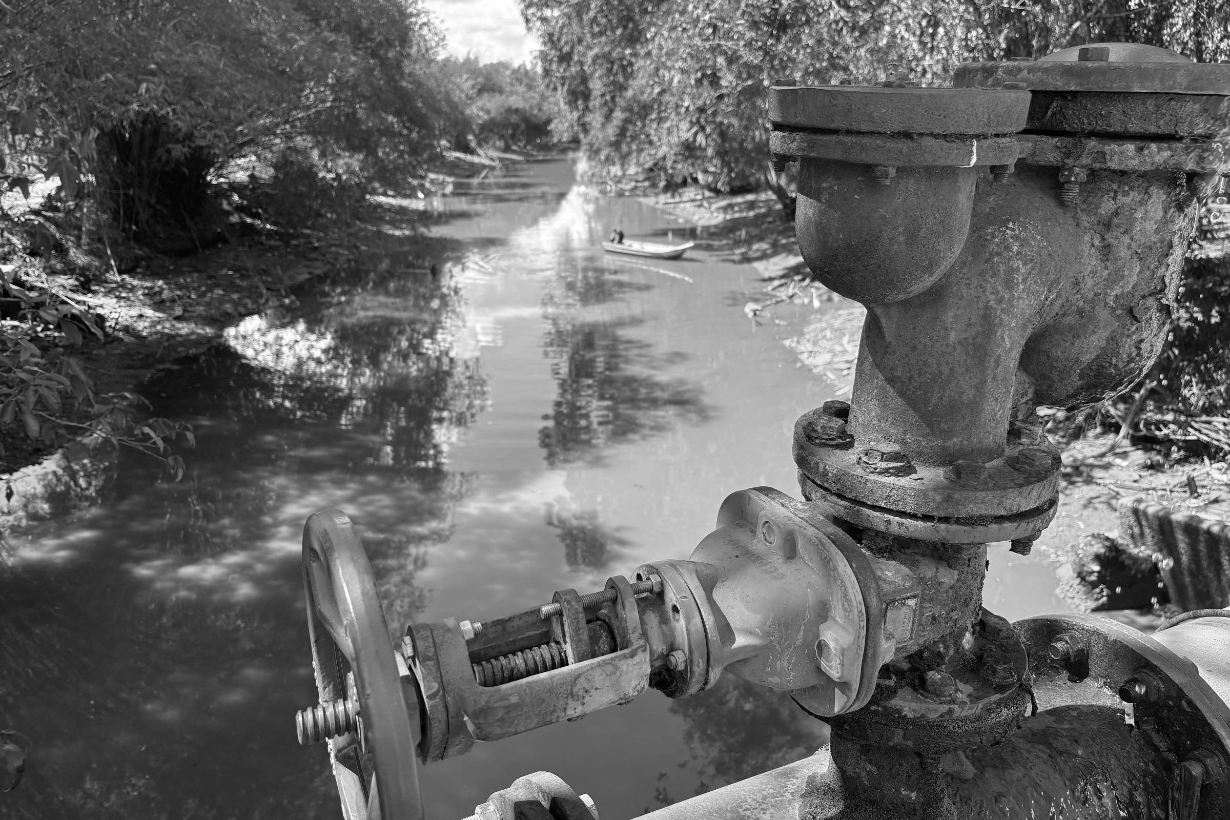 A imagem mostra uma válvula de metal enferrujada em primeiro plano, com uma roda azul para controle. Ao fundo, há um rio com água clara e vegetação nas margens, refletindo a luz do sol. A cena é tranquila, com árvores ao redor e um céu parcialmente nublado.