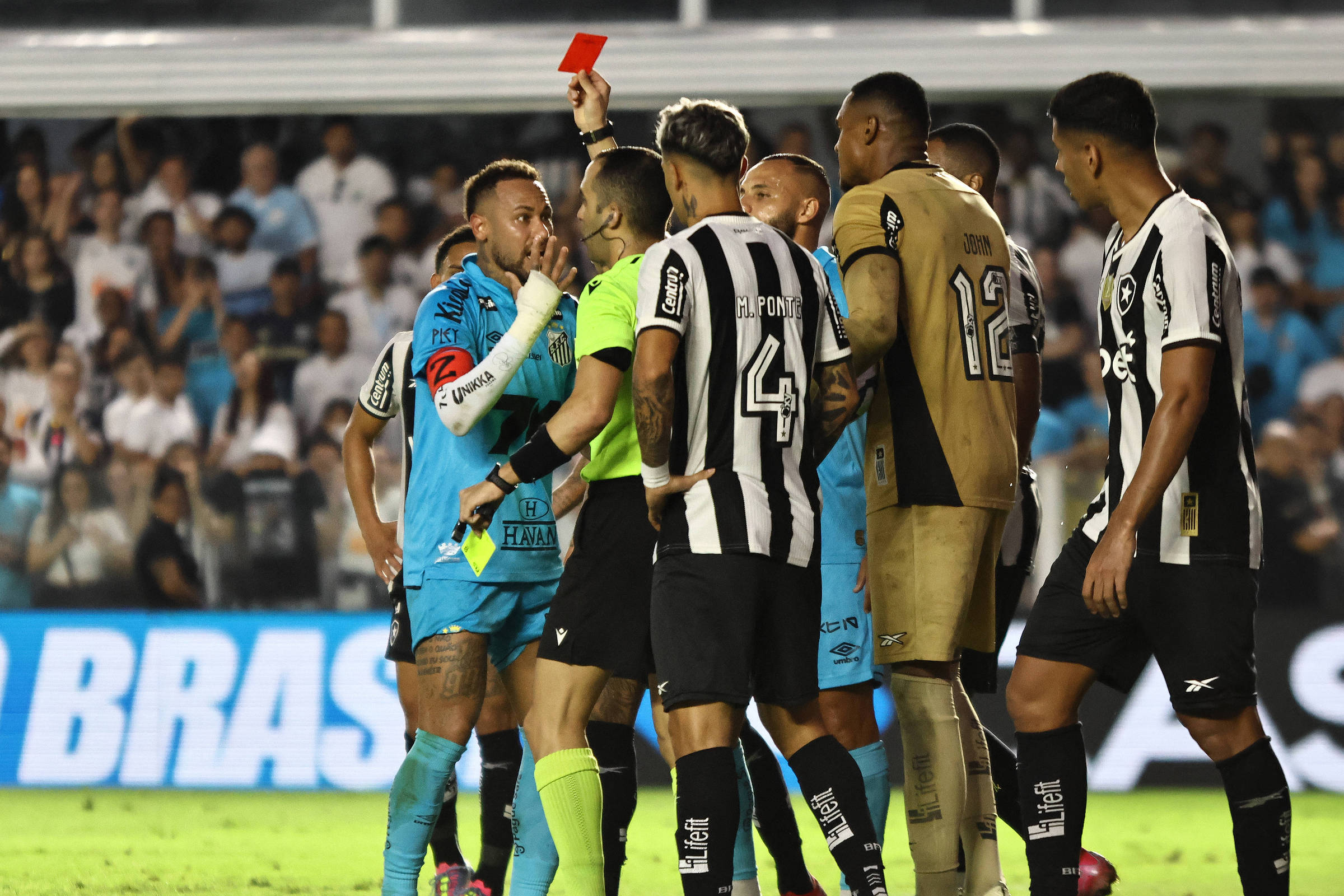 A imagem mostra um momento de tensão em um jogo de futebol, onde um árbitro está mostrando um cartão vermelho para um jogador. O jogador, que está usando um uniforme azul, parece estar discutindo com o árbitro, que está vestido de amarelo. Outros jogadores, de diferentes uniformes, estão ao redor, observando a situação. O fundo da imagem mostra uma multidão de torcedores.