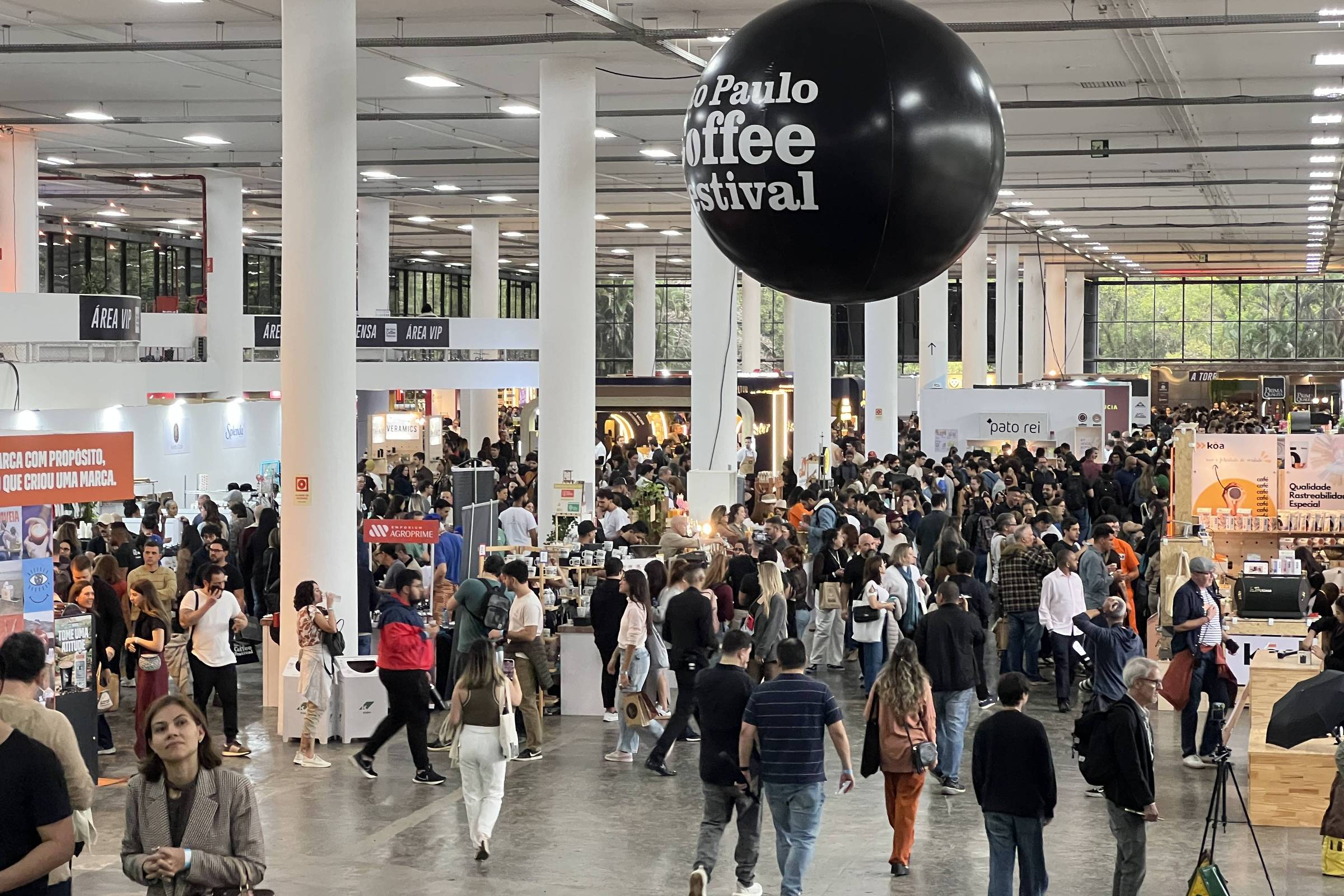 A imagem mostra uma grande multidão em um evento, possivelmente um festival de café, com várias barracas e estandes ao fundo. Um balão preto com a inscrição