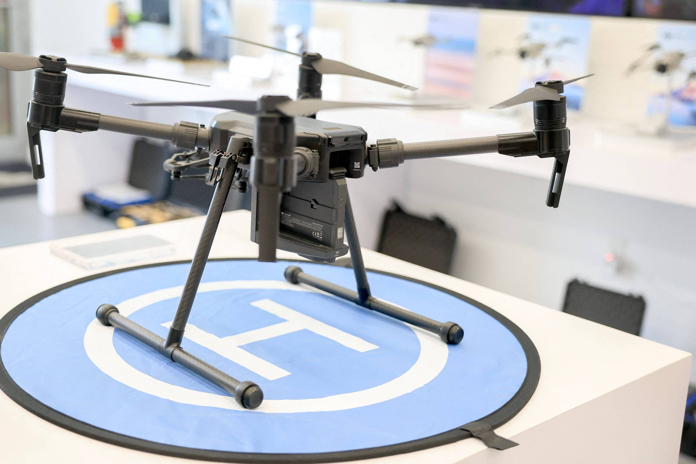 Um drone preto está posicionado em cima de um círculo azul com um símbolo de pouso. O drone possui quatro hélices e uma estrutura robusta, com uma câmera visível na parte frontal. Ao fundo, há uma parede com várias imagens e equipamentos relacionados a drones.