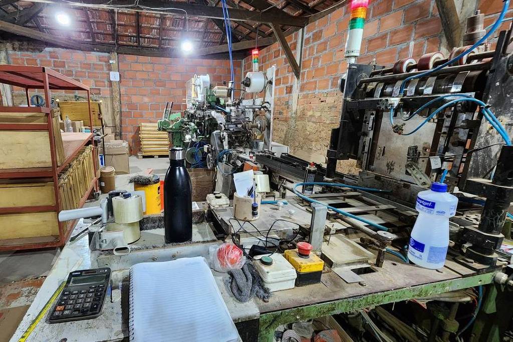 A imagem mostra um ambiente de trabalho com várias máquinas e equipamentos. À esquerda, há uma mesa de trabalho com uma calculadora, um caderno em branco, um frasco de líquido e um recipiente preto. Ao fundo, várias caixas de madeira estão empilhadas. No centro, uma máquina complexa está visível, com tubos e fios expostos. O teto é de madeira e as paredes são de tijolos expostos. Há também luzes indicadoras coloridas no topo da máquina