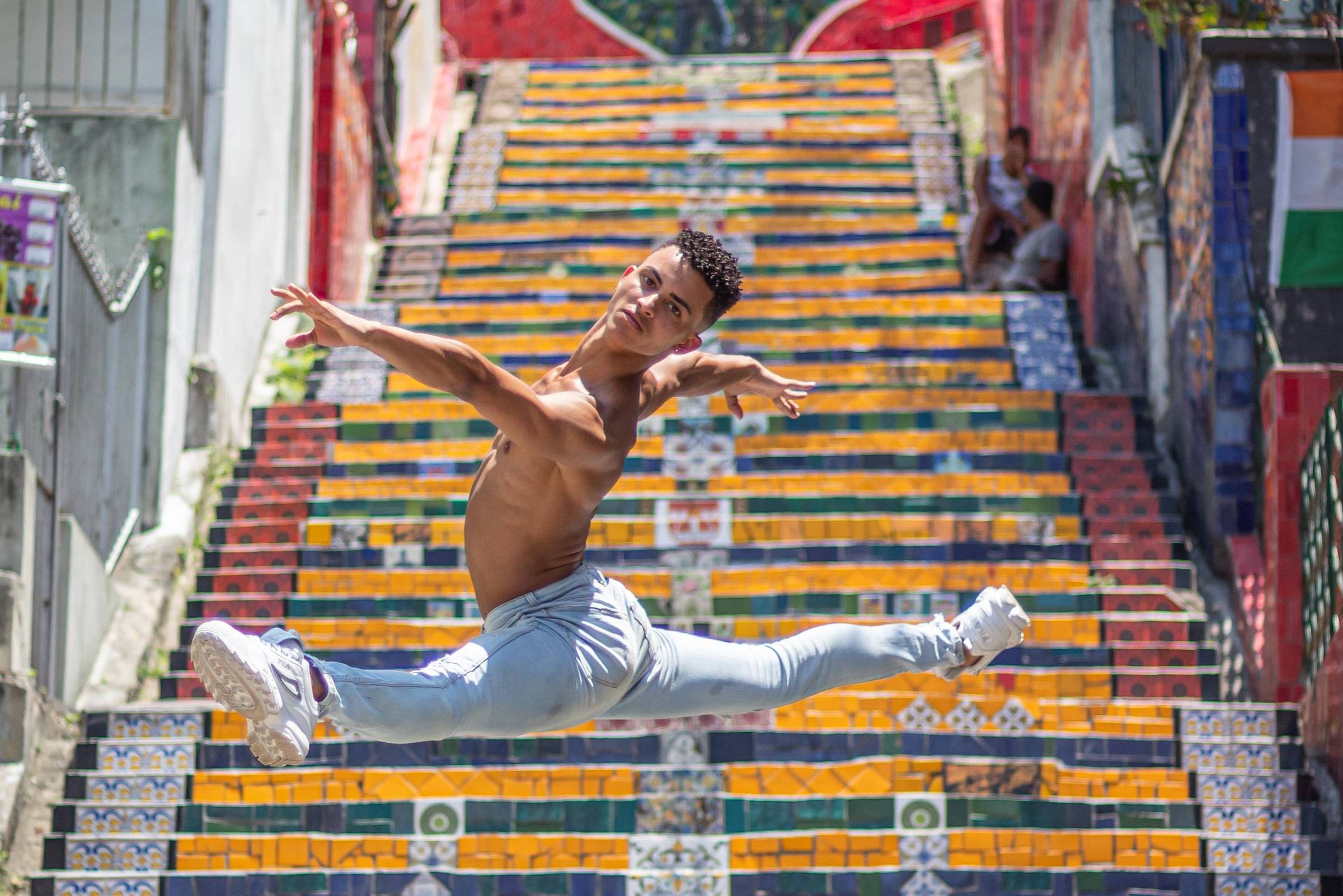 Um dançarino está realizando um salto acrobático em uma escadaria decorada com azulejos coloridos. A escadaria apresenta uma variedade de cores e padrões, com destaque para a palavra