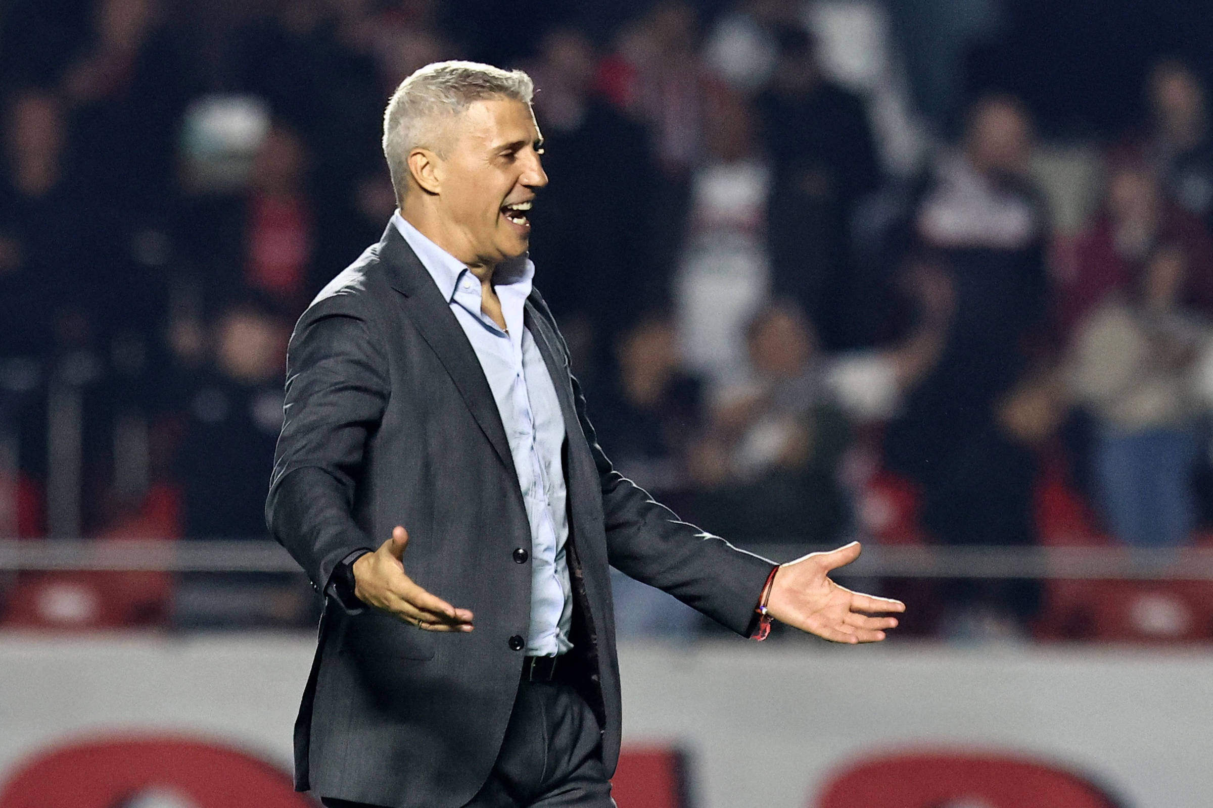 Um homem de cabelo curto e grisalho, vestido com um terno escuro e uma camisa clara, está em um campo de futebol. Ele parece estar animado e gesticulando com as mãos, enquanto observa a partida. Ao fundo, há uma multidão de torcedores, com algumas pessoas visíveis nas arquibancadas.
