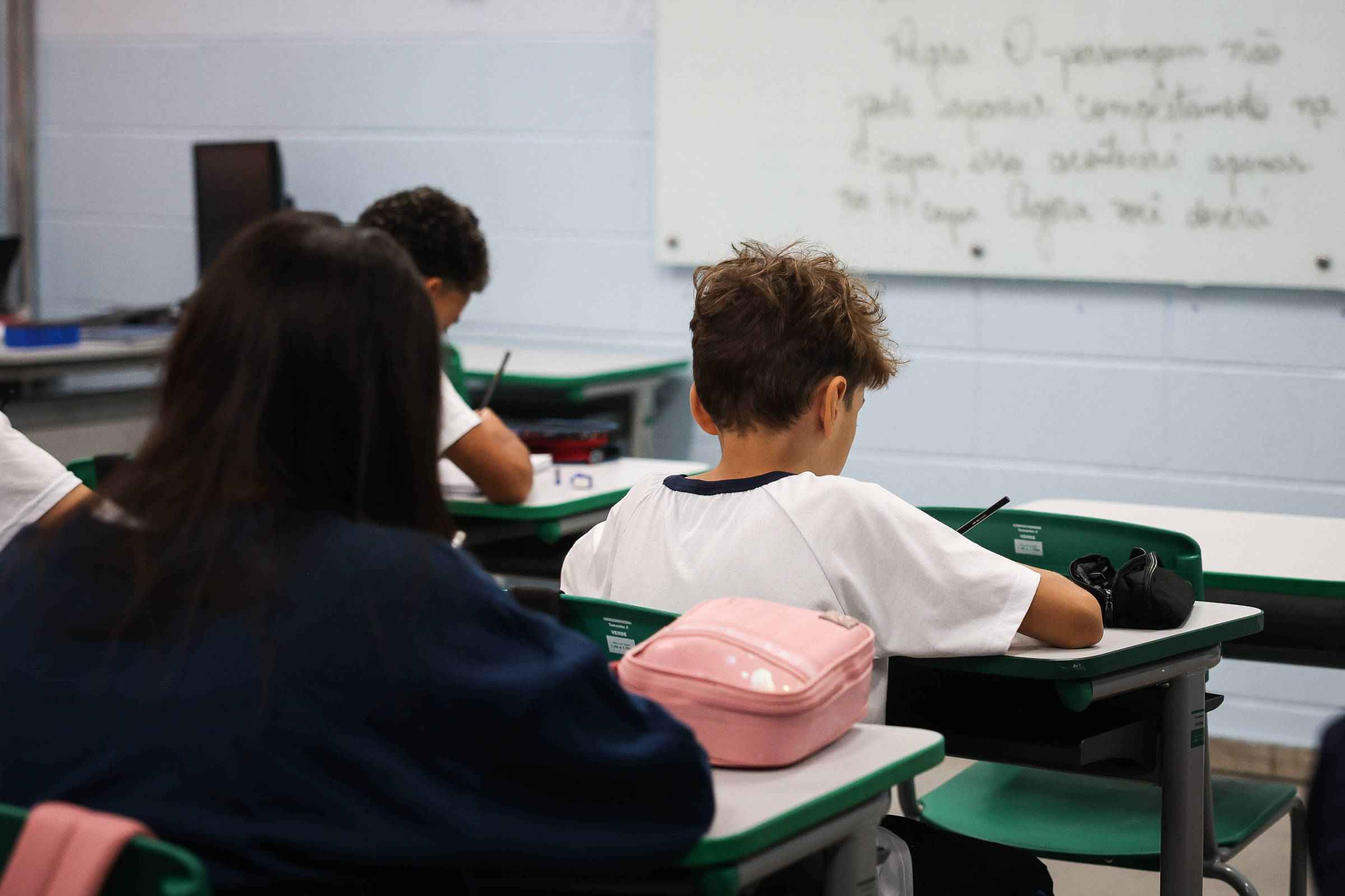 A imagem mostra uma sala de aula com estudantes sentados em carteiras. Um menino de cabelo curto, usando uma camiseta branca, está concentrado escrevendo em um caderno. À sua frente, há uma mochila rosa e uma câmera. Ao fundo, uma lousa branca com texto escrito. Outros alunos também estão visíveis, mas apenas a parte de trás de suas cabeças é visível.