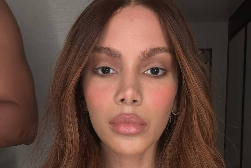 A imagem mostra um retrato de uma mulher com cabelo longo e castanho claro. Ela está usando uma blusa sem mangas de cor clara e colares delicados. O fundo é neutro, com uma parede e uma parte de um braço visível à esquerda. A mulher tem uma expressão neutra e está olhando diretamente para a câmera.