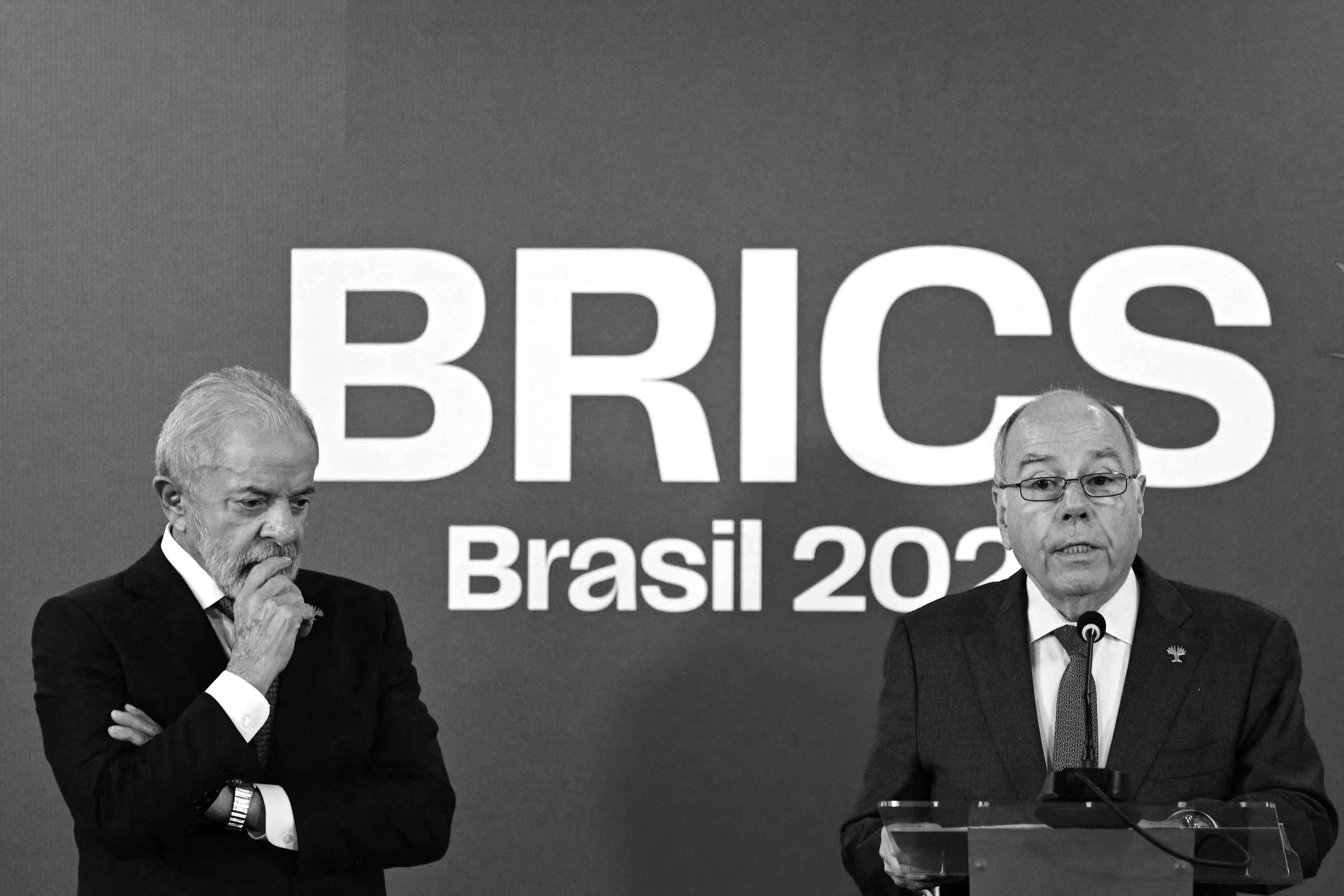 A imagem mostra dois homens em um evento relacionado ao BRICS. Um deles está à esquerda, com cabelo grisalho e barba, vestindo um paletó escuro e com uma expressão pensativa. O outro homem, à direita, está falando ao microfone, usando óculos e um paletó claro. Ao fundo, há um painel azul com a inscrição