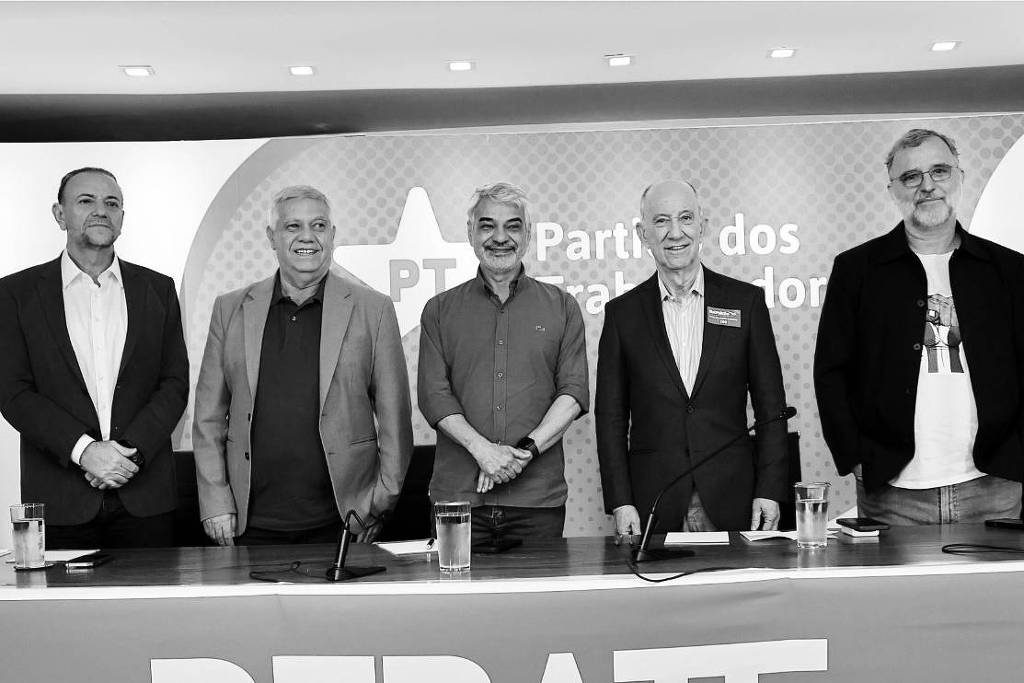 A imagem mostra um grupo de seis pessoas em um evento de debate, posando em uma mesa. Todos estão em pé, com expressões sérias. A mesa tem copos de água e um pano com a palavra