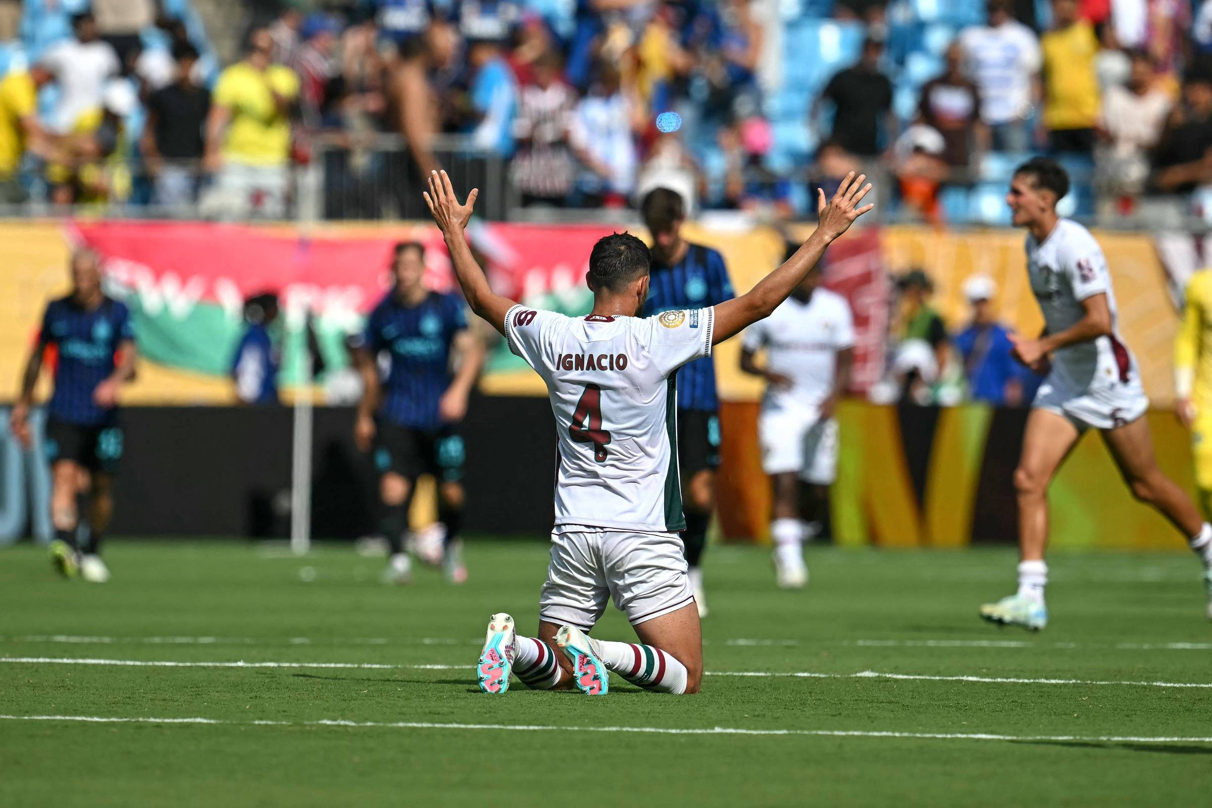 Um jogador de futebol está ajoelhado no campo, com as mãos levantadas em celebração. Ele usa um uniforme branco com detalhes em vermelho e o número 7 nas costas. Ao fundo, outros jogadores estão em movimento, e há uma multidão nas arquibancadas. O clima parece ser de comemoração após um gol.