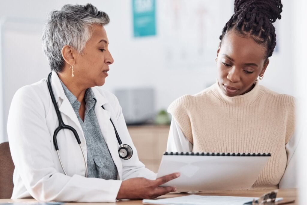 Jovem no consultório com médica analisando exames. A médica tem o cabelo curto e grisalho e está usando jaleco branco e camisa cinza e com estetoscópio no pescoço e a paciente tem o cabelo trançado preso em coque e está usando camisa de manga longa branca e colete bege.