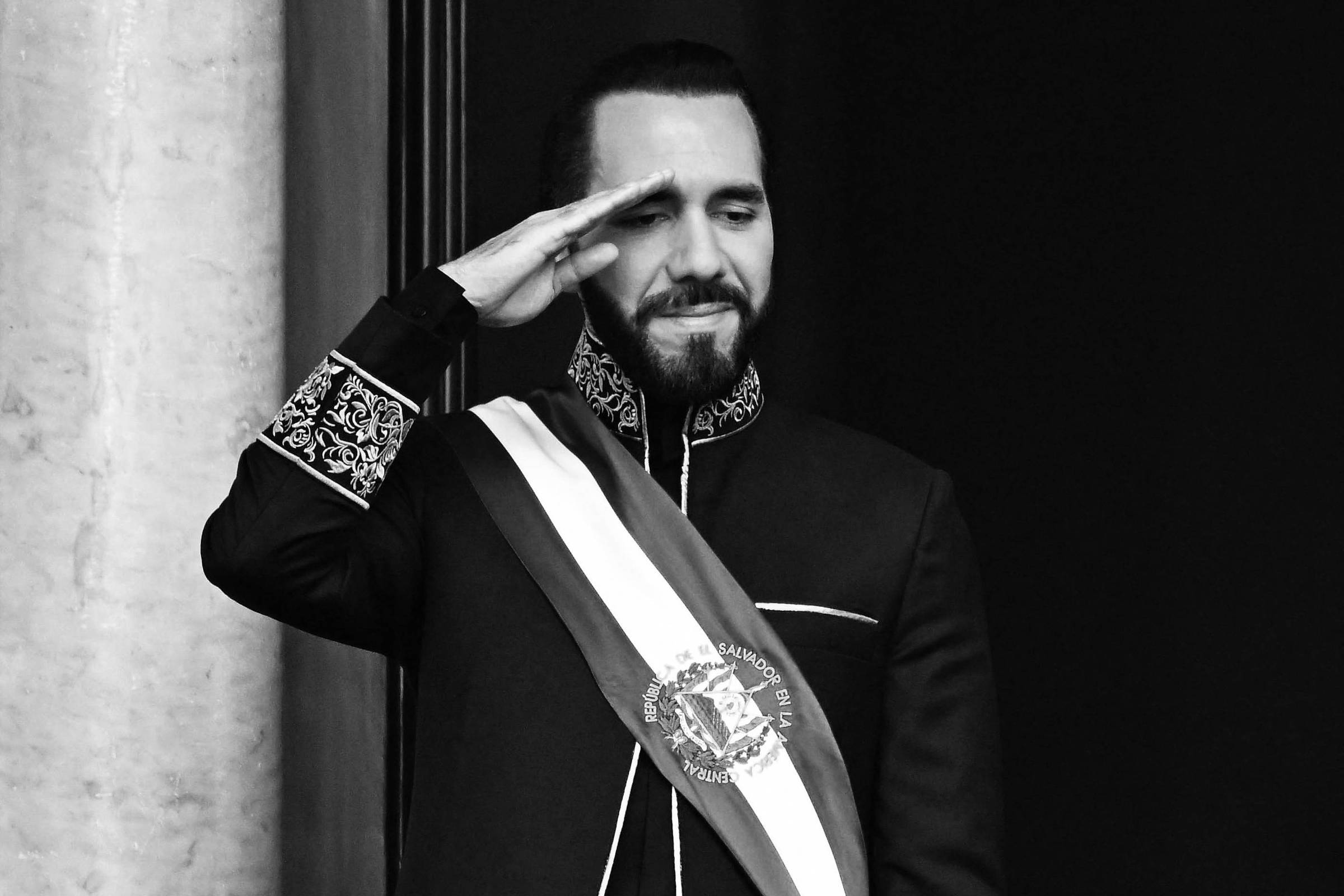 Um homem em um traje formal escuro, com detalhes em dourado, está fazendo um gesto de saudação. Ele usa uma faixa azul e branca com um emblema no centro. O fundo é de uma parede clara, e ele parece estar em uma posição elevada, possivelmente em um balcão.