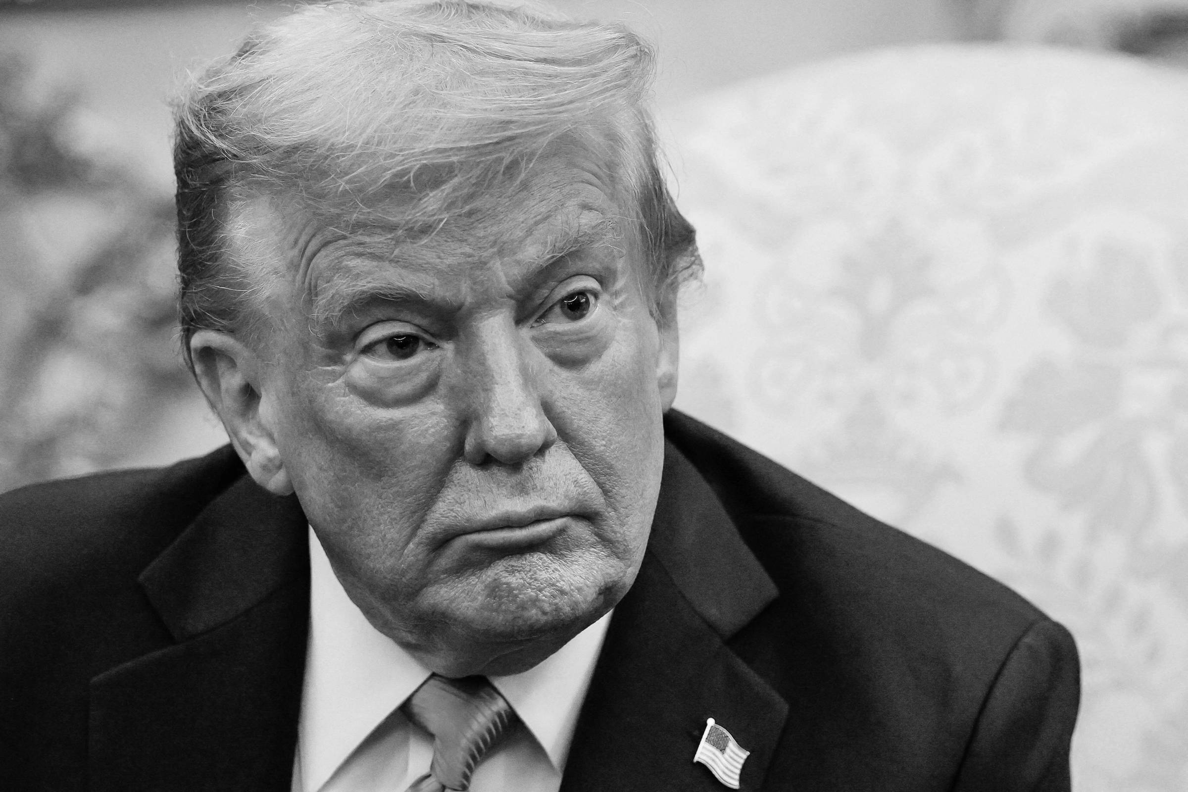 Um homem com cabelo loiro e pele bronzeada está sentado em um ambiente formal. Ele usa um terno escuro e uma gravata vermelha. O fundo apresenta um padrão claro e detalhes decorativos em dourado. O homem tem uma expressão séria e está olhando para frente.