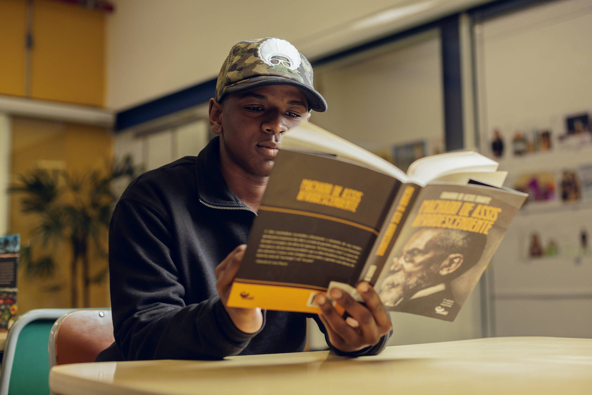 Um jovem está sentado à mesa em um ambiente interno, lendo um livro intitulado