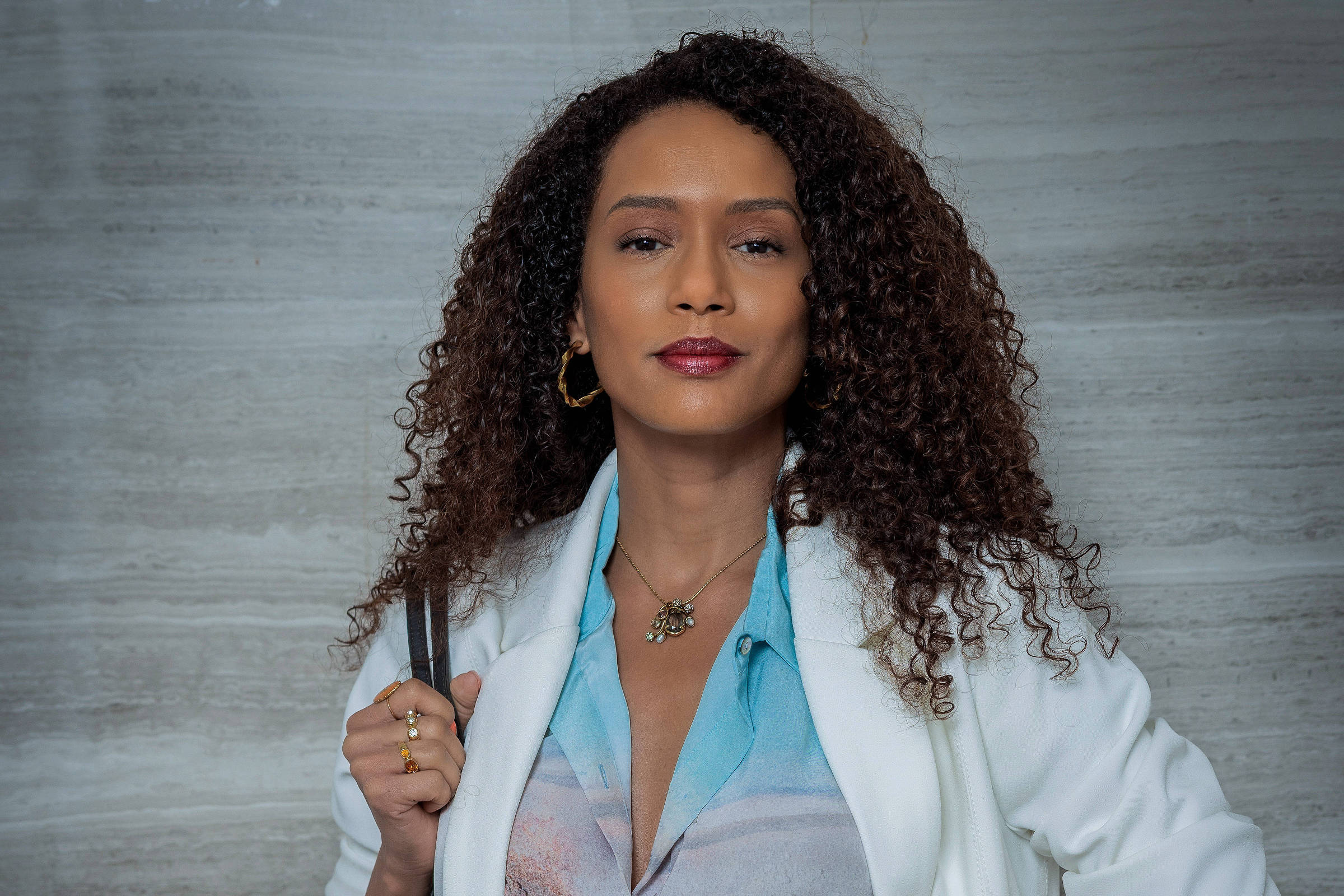 A imagem mostra uma mulher com cabelo cacheado e longo, vestindo um blazer branco sobre uma blusa colorida e calças brancas. Ela está segurando uma bolsa grande e escura, e usa acessórios como brincos e um relógio. O fundo é uma parede de textura clara.