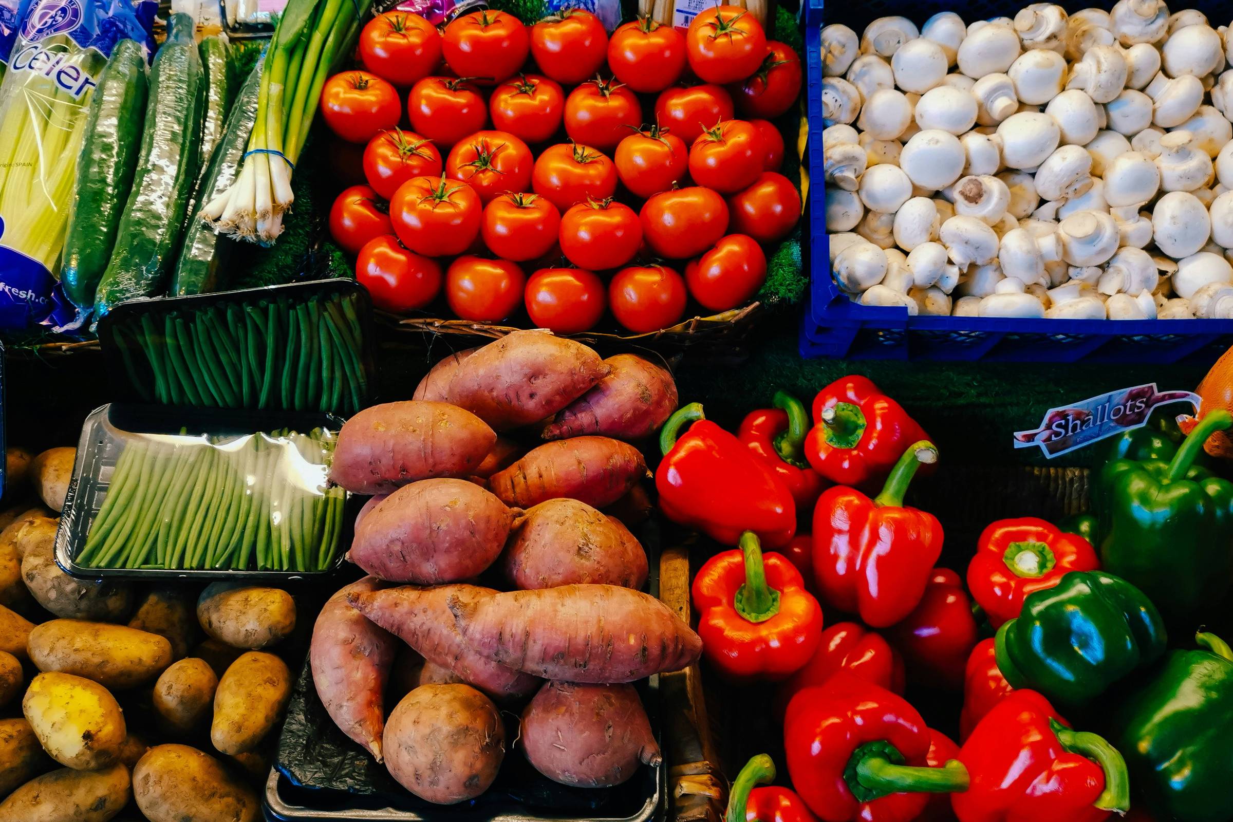 Imagem de uma banca de vegetais em um mercado. Há uma variedade de vegetais, incluindo tomates, cogumelos, pimentões vermelhos e verdes, batatas, batatas-doces, feijões verdes, cebolinhas, pepinos embalados e outros vegetais embalados.