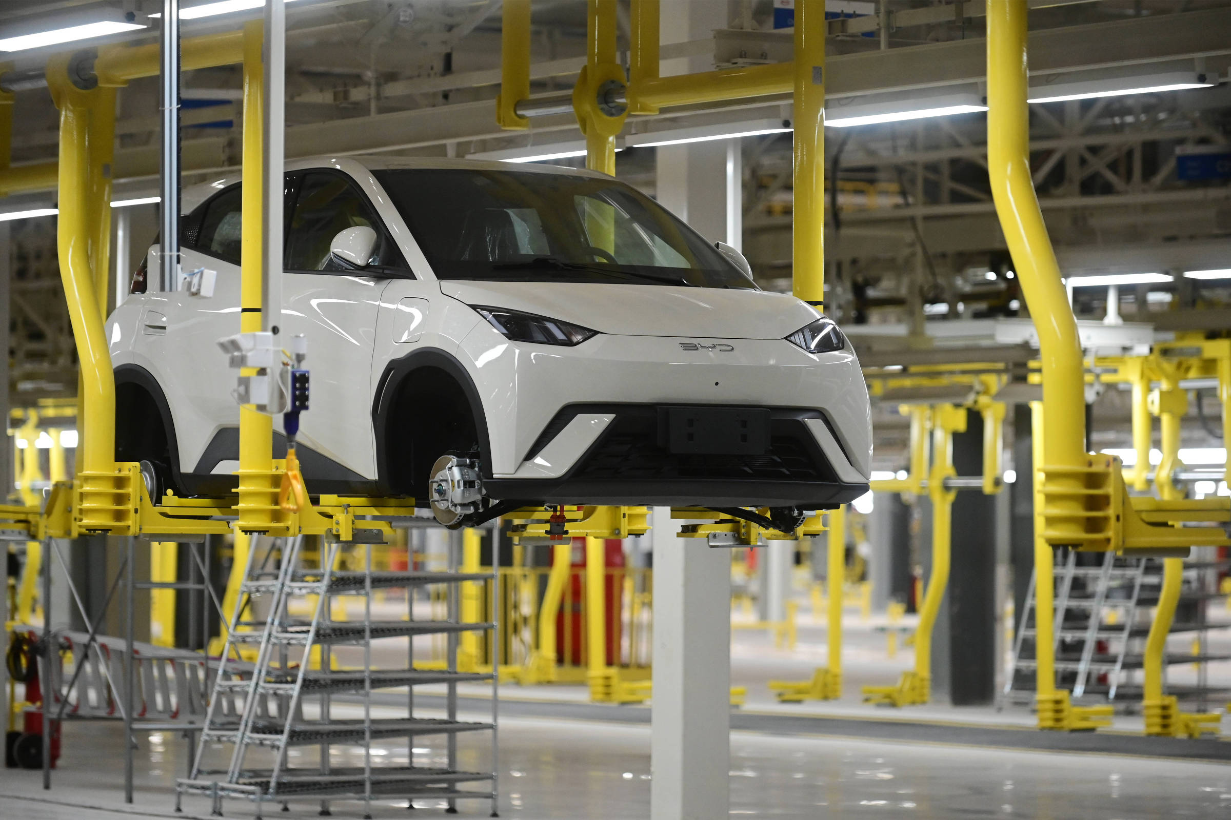 A imagem mostra um carro branco suspenso em uma linha de montagem. O veículo está sem rodas e é apoiado por estruturas amarelas. O ambiente é uma fábrica com iluminação fluorescente e piso claro, com equipamentos e ferramentas visíveis ao fundo.