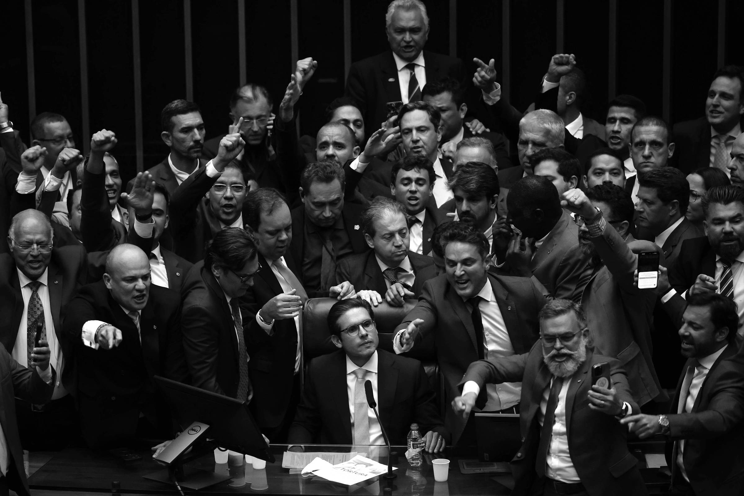 A imagem mostra um grupo de parlamentares em uma sessão do Congresso Nacional. Eles estão em pé, com expressões intensas, alguns levantando os punhos e outros gesticulando. O ambiente é formal, com mesas e equipamentos de votação ao fundo. A maioria dos homens está vestida com ternos e gravatas, e há uma sensação de agitação e debate acalorado entre os presentes.