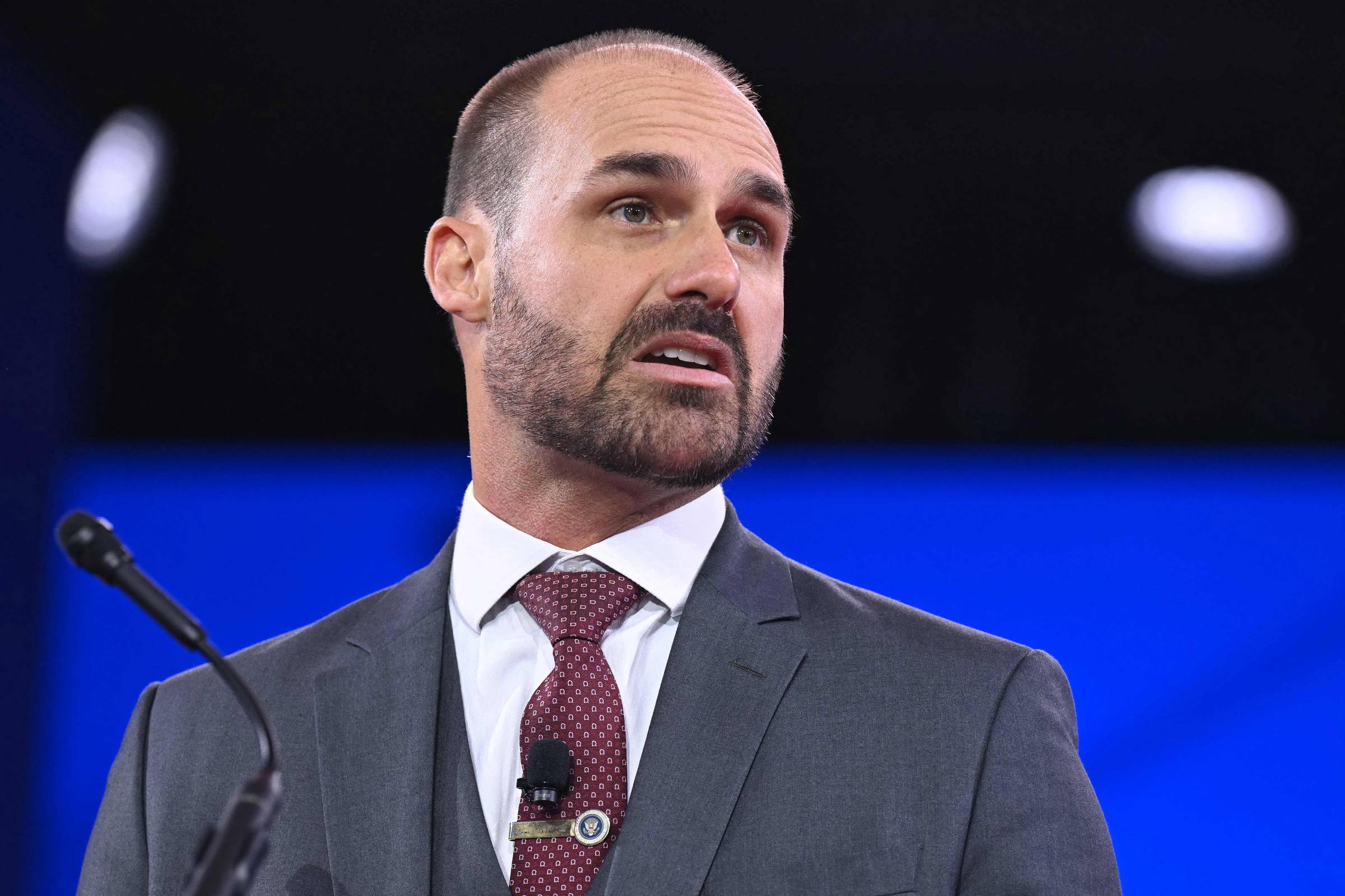 Um homem com cabelo curto e barba está falando em um microfone. Ele usa um terno cinza e uma gravata vermelha com padrões. O fundo é desfocado, mas parece ser um ambiente de conferência ou evento. O homem parece estar expressando uma ideia ou opinião de forma enfática.