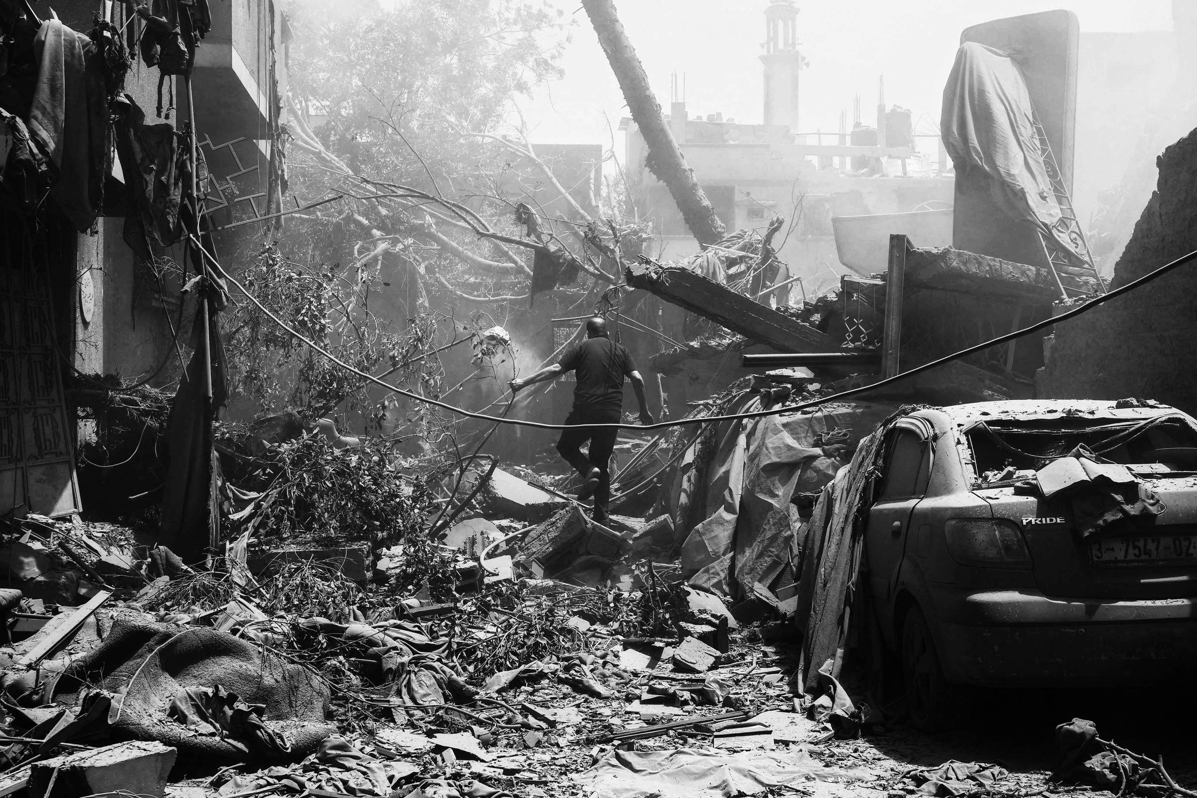 A imagem mostra um cenário de destruição urbana, com escombros e destroços visíveis em um ambiente empoeirado. Um homem caminha entre os destroços, que incluem fios elétricos caídos e um carro danificado coberto de poeira. Ao fundo, há edifícios danificados e uma atmosfera de desolação.