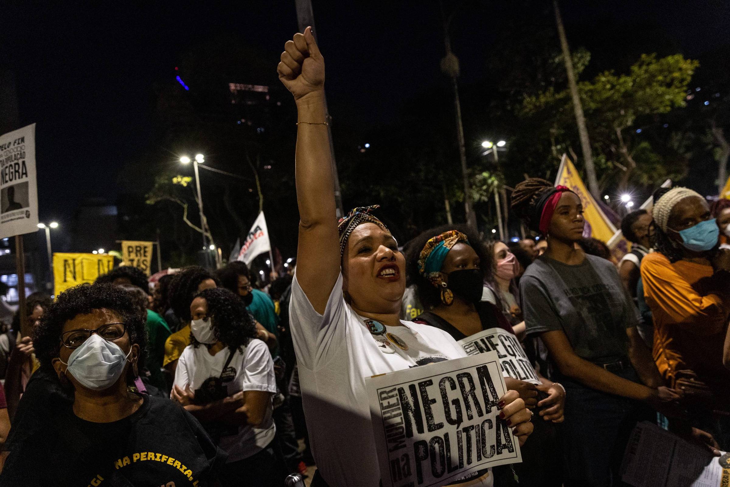 A imagem mostra um grupo de pessoas participando de um protesto à noite. No primeiro plano, uma mulher levanta a mão com um cartaz que diz