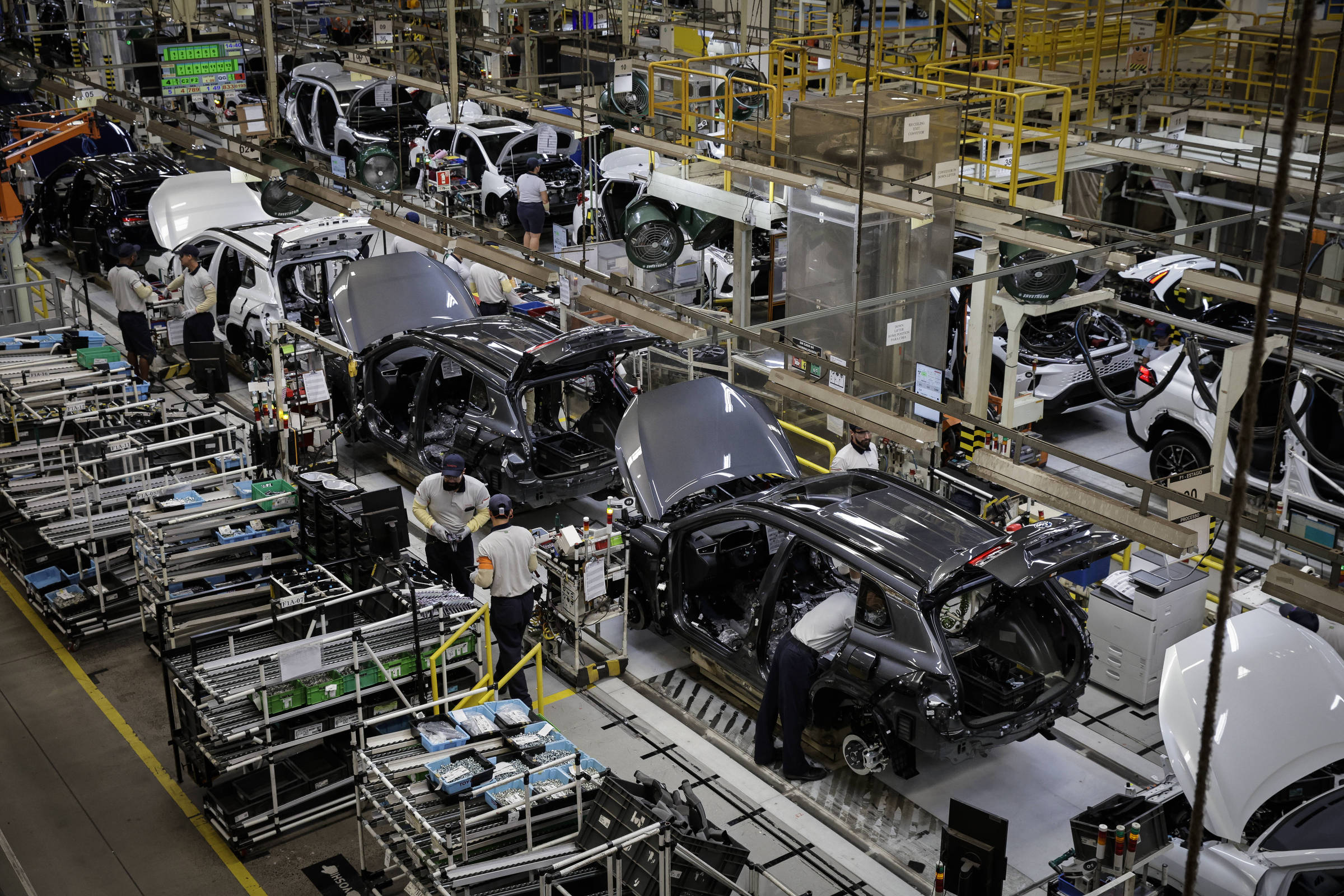 A imagem mostra uma linha de montagem em uma fábrica de automóveis. Vários carros em diferentes estágios de montagem estão alinhados, com trabalhadores realizando tarefas em cada veículo. O ambiente é industrial, com equipamentos e ferramentas visíveis ao redor. Há também estruturas metálicas e prateleiras com peças e componentes.