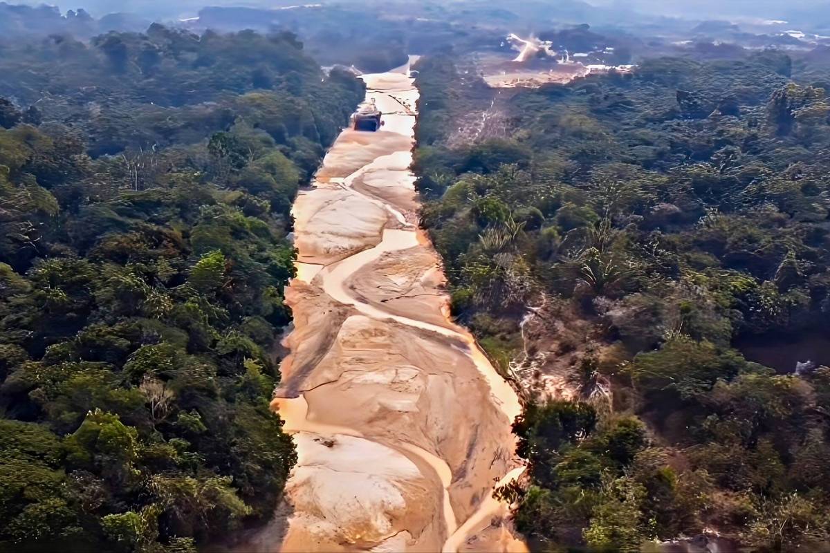 A imagem mostra um caminho sinuoso em uma área florestal, com solo exposto e vegetação densa ao redor. O caminho é de terra e areia, e há uma máquina visível ao fundo, sugerindo atividade de extração ou construção. A paisagem é cercada por árvores e a atmosfera parece neblinada.