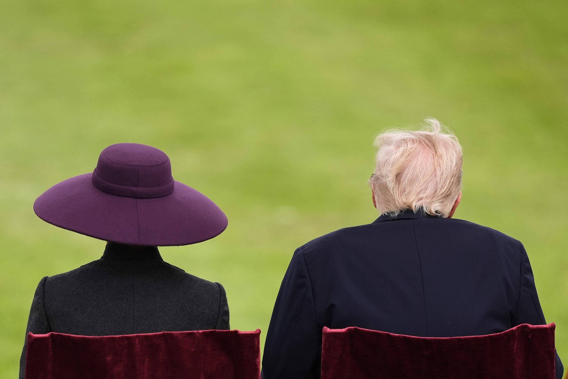 A imagem mostra as costas de duas pessoas sentadas em cadeiras vermelhas. A pessoa à esquerda usa um chapéu roxo grande e um casaco escuro, enquanto a pessoa à direita tem cabelo loiro e está vestindo um paletó escuro. O fundo é um gramado verde.