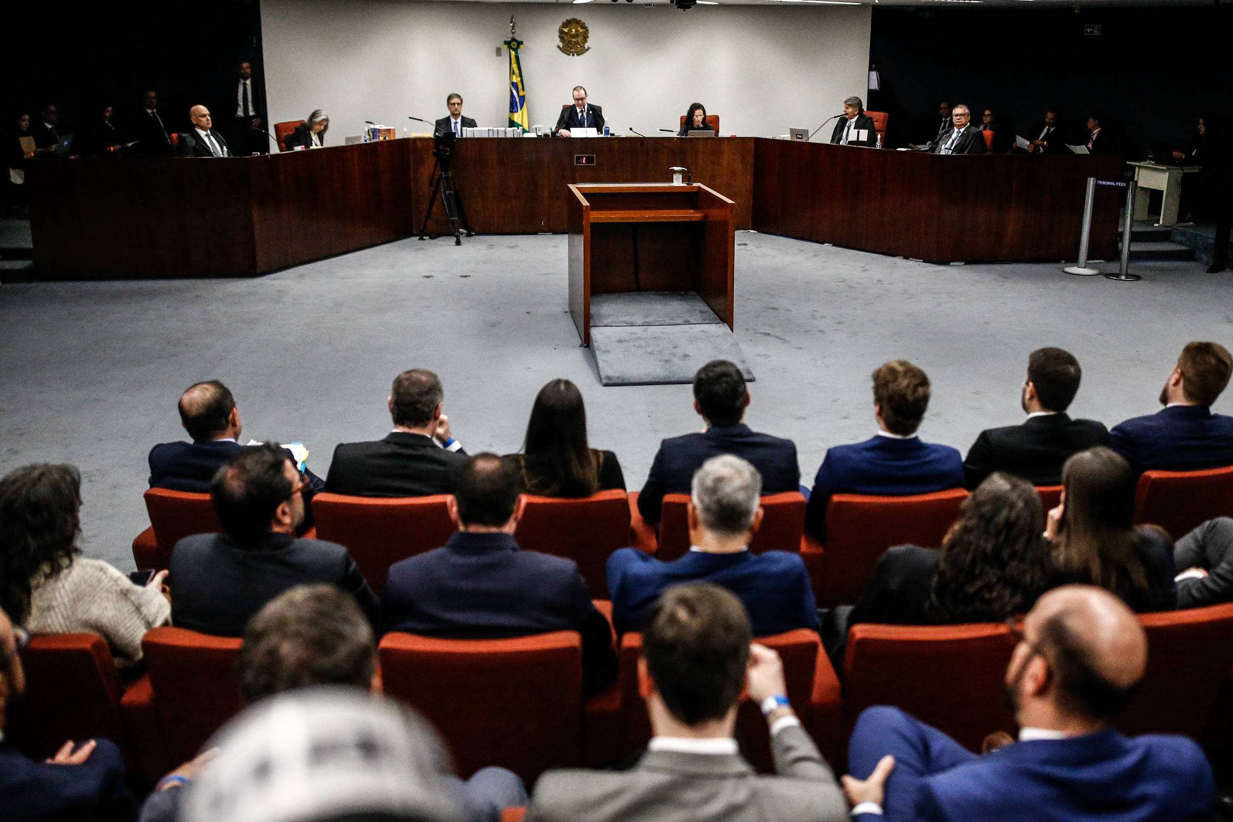 A imagem mostra uma audiência em um tribunal. No centro, há uma mesa de audiência com um microfone e um copo de água. Atrás da mesa, estão sentados vários juízes e advogados, enquanto na frente, um grupo de pessoas está assistindo, algumas em trajes formais. Ao fundo, há uma bandeira do Brasil e o brasão nacional. O ambiente é bem iluminado e possui um design moderno.
