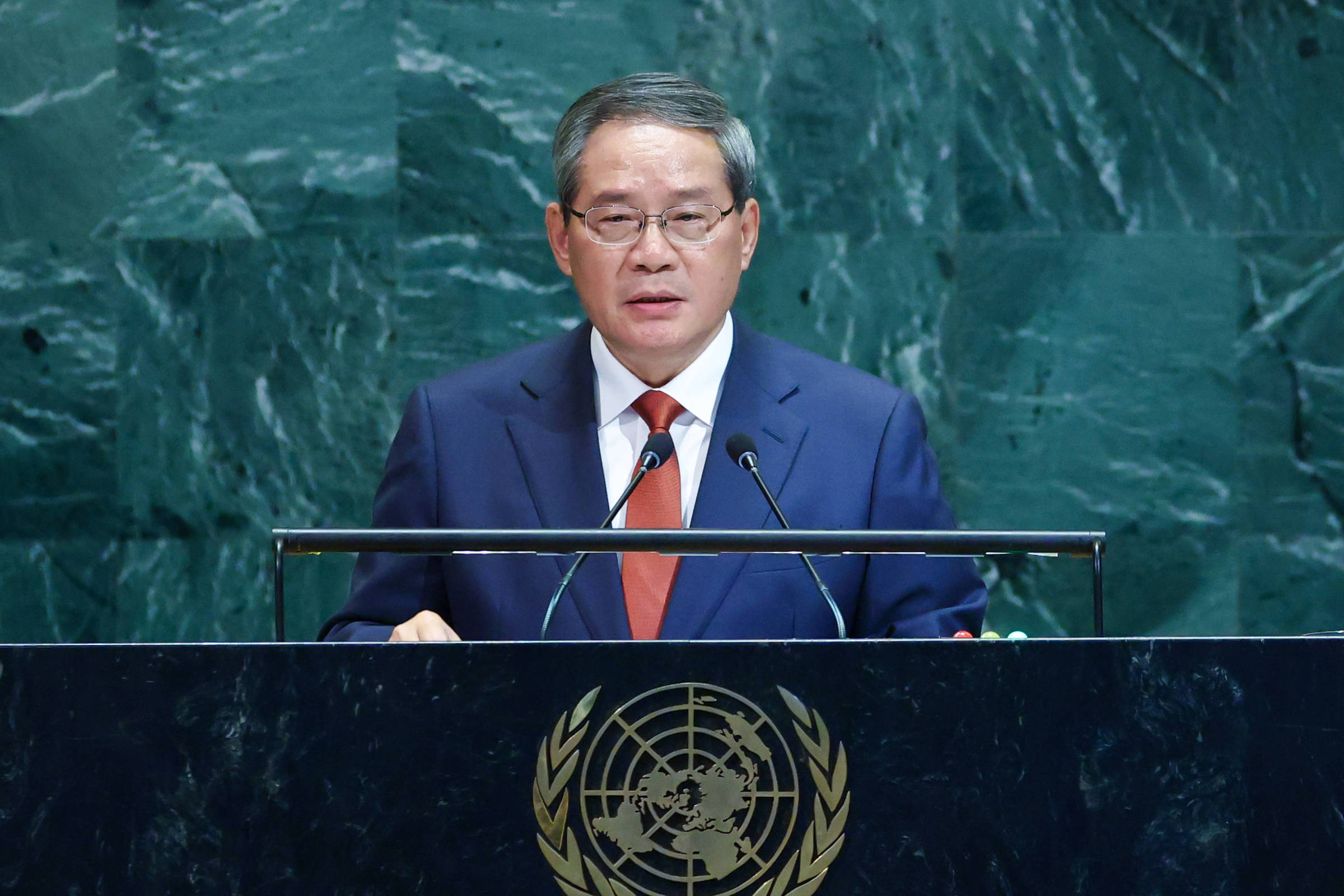 Homem de terno azul e gravata vermelha fala em púlpito com símbolo da ONU. Fundo de mármore verde escuro na Assembleia Geral.