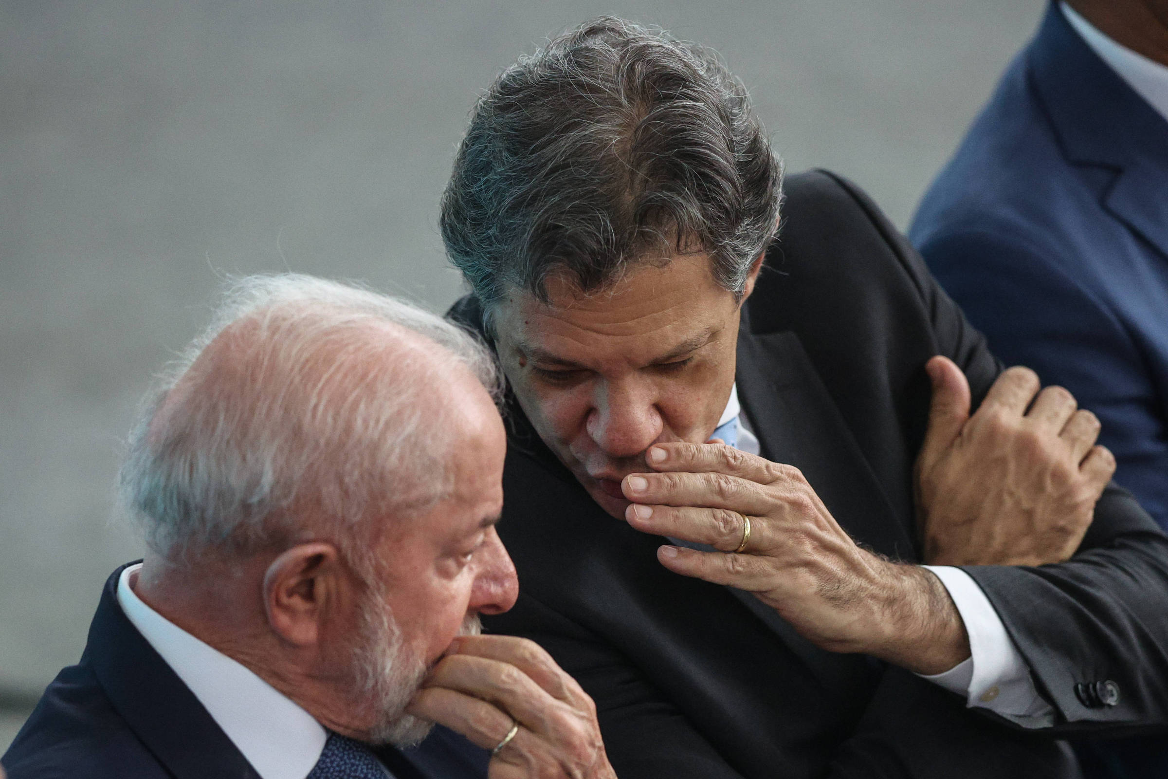 A imagem mostra dois homens sentados em um evento. O homem à esquerda tem cabelo grisalho e uma barba branca, vestindo um terno escuro e uma gravata. O homem à direita tem cabelo castanho e está vestido com um terno claro. Eles estão inclinados um em direção ao outro, parecendo conversar em um tom reservado.