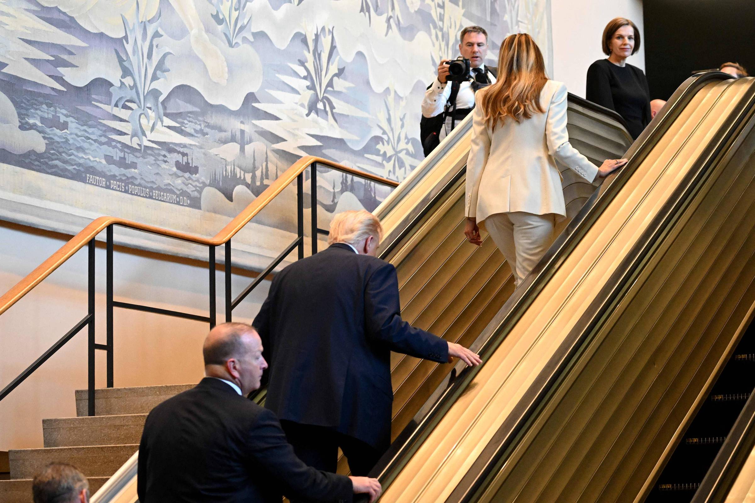 A imagem mostra um grupo de pessoas subindo uma escada rolante. Um homem de terno escuro está subindo a escada, seguido por uma mulher vestindo um terno claro. Ao fundo, há uma parede decorada com um mural que apresenta um cenário com elementos naturais. Algumas pessoas estão observando e uma delas está segurando uma câmera, possivelmente tirando fotos. O ambiente parece ser um espaço público ou institucional.