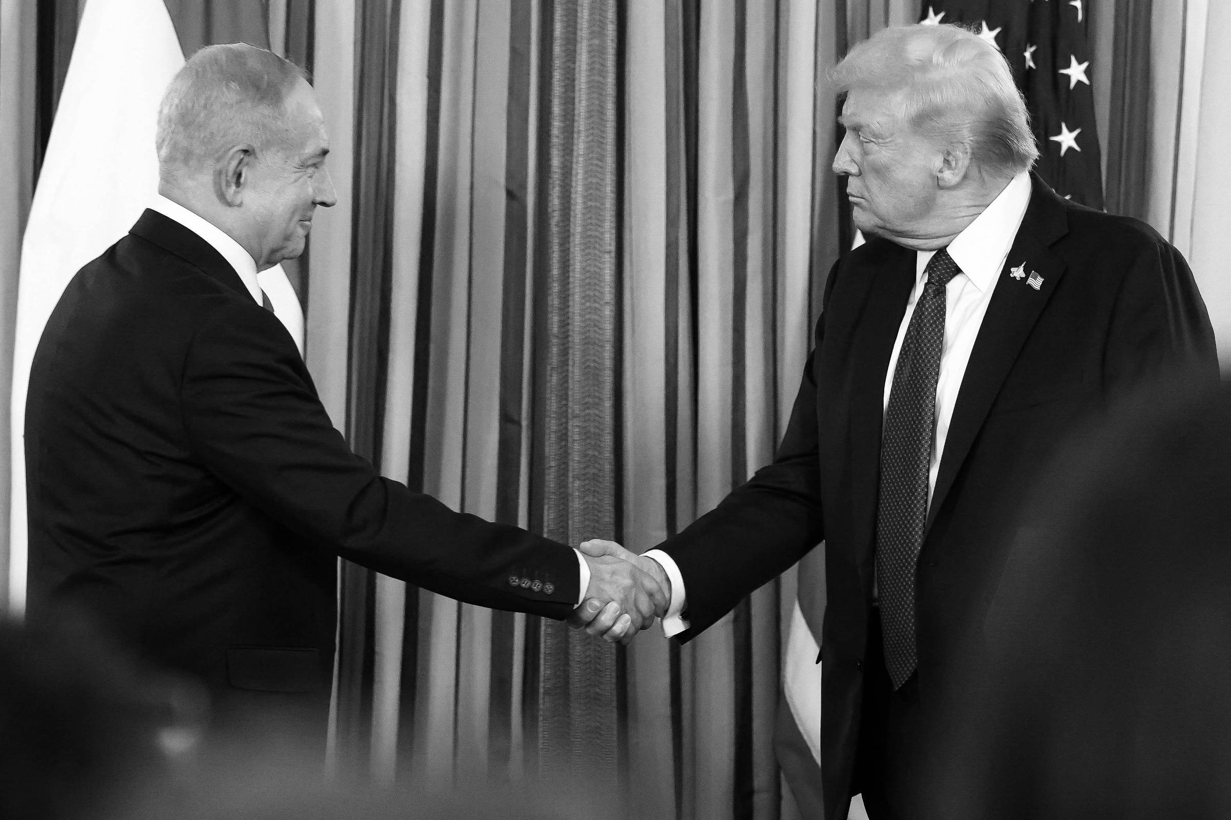 Benjamin Netanyahu e Donald Trump apertam as mãos em ambiente formal com bandeiras de Israel e Estados Unidos ao fundo.