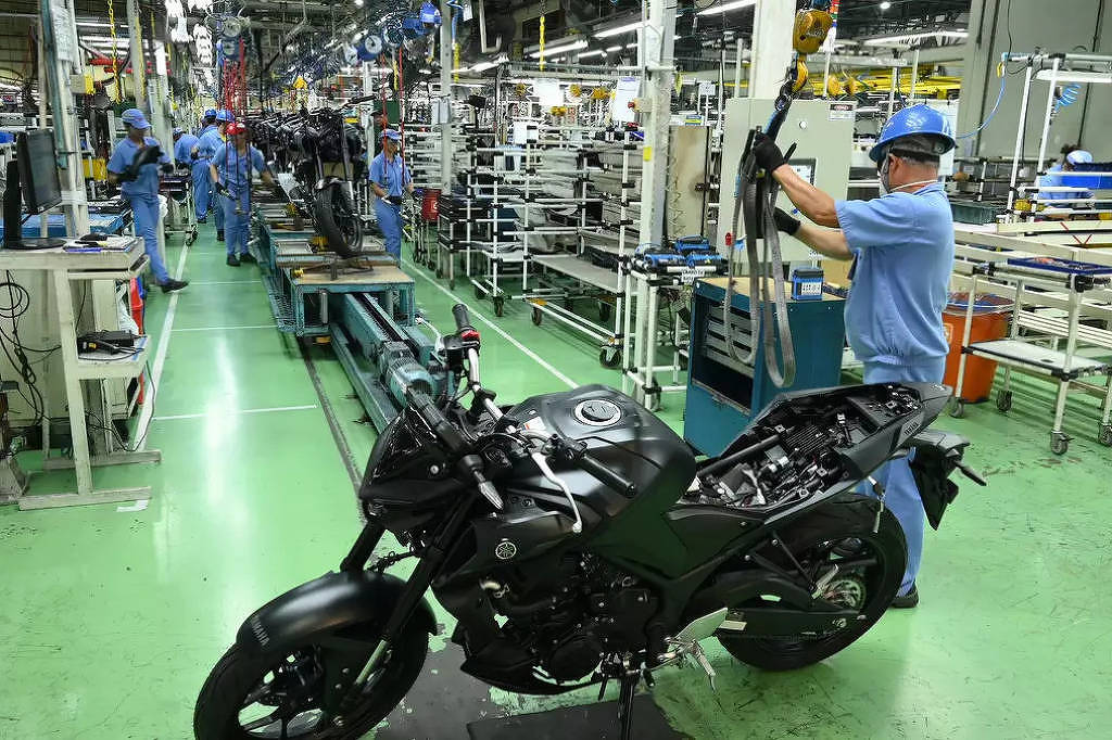 A imagem mostra uma linha de montagem de motocicletas em uma fábrica. No primeiro plano, há uma motocicleta preta parcialmente montada. Ao fundo, vários trabalhadores em uniformes azuis estão envolvidos na montagem de outras motocicletas, com ferramentas e peças ao redor. O ambiente é bem iluminado e organizado, com prateleiras e equipamentos de montagem visíveis.