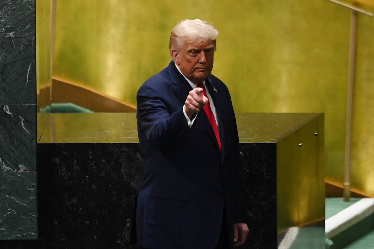 Um homem com cabelo loiro e terno escuro está em um palco, apontando com o dedo indicador. O fundo é de uma parede verde e há uma escada ao lado. O homem parece estar em um momento de discurso ou apresentação.