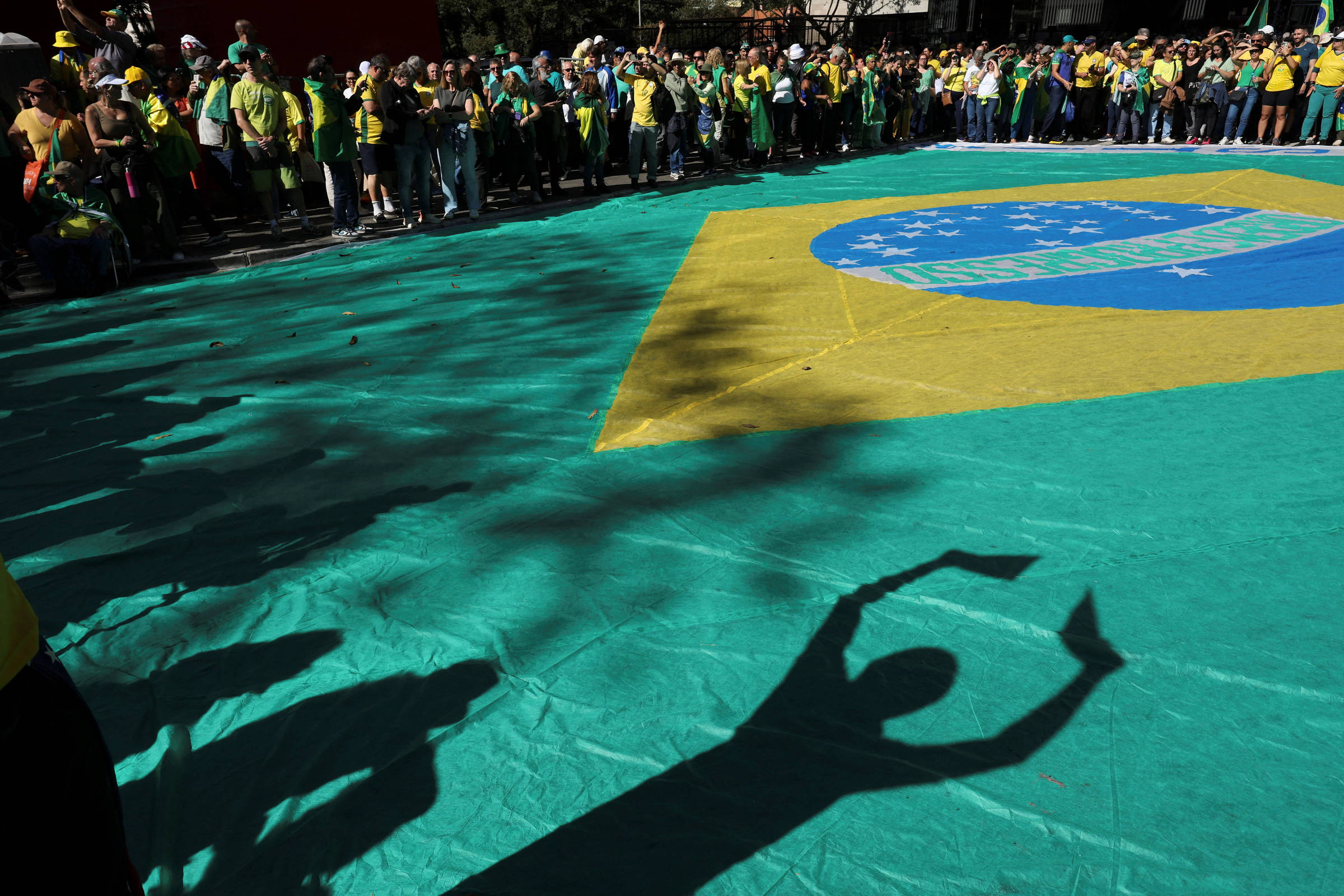 A imagem mostra uma grande bandeira do Brasil estendida no chão, com pessoas ao redor vestindo roupas nas cores verde e amarelo. A sombra de uma mão é projetada sobre a bandeira, formando um gesto de