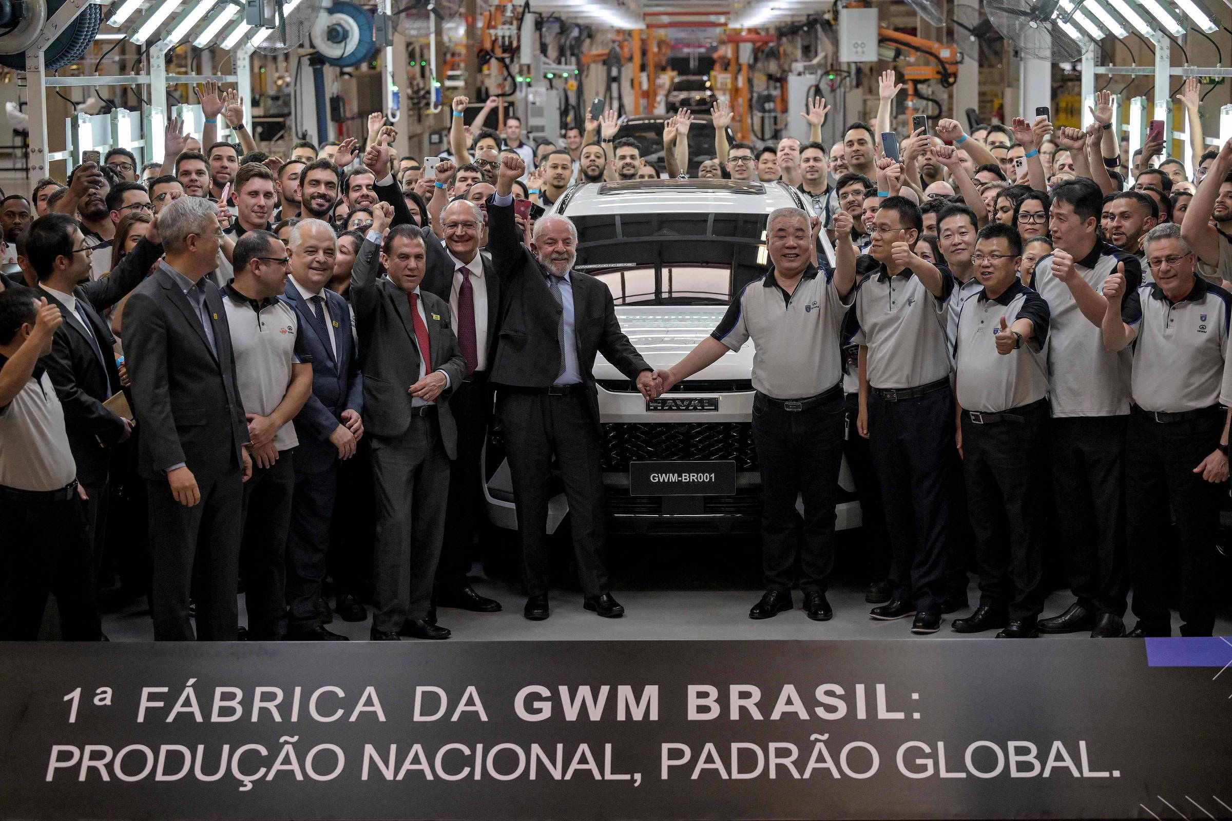 A imagem mostra um grupo grande de pessoas reunidas em uma fábrica, celebrando a inauguração da primeira fábrica da GWM Brasil. No centro, algumas pessoas estão segurando as mãos, enquanto outras estão levantando os braços em sinal de comemoração. Ao fundo, é possível ver um veículo da marca GWM e uma linha de produção. Na parte inferior da imagem, há um banner com a frase: