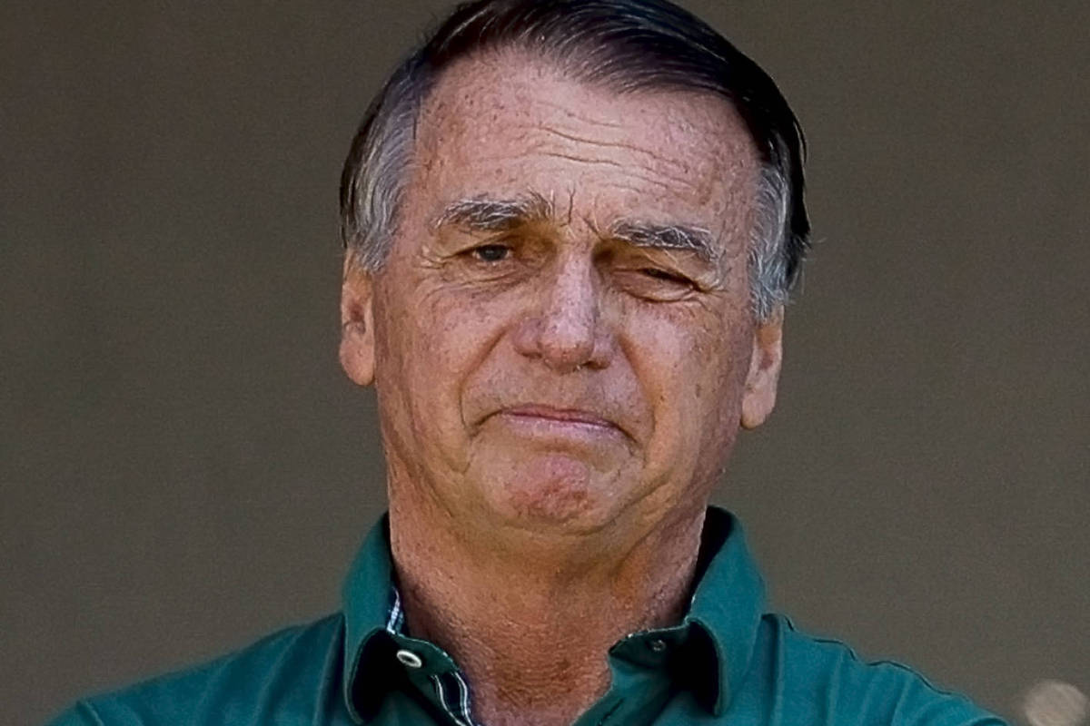Um homem de pele clara, com cabelo grisalho e uma expressão séria. Ele está usando uma camisa verde e tem os braços cruzados. O fundo é neutro e desfocado, destacando o homem na imagem.