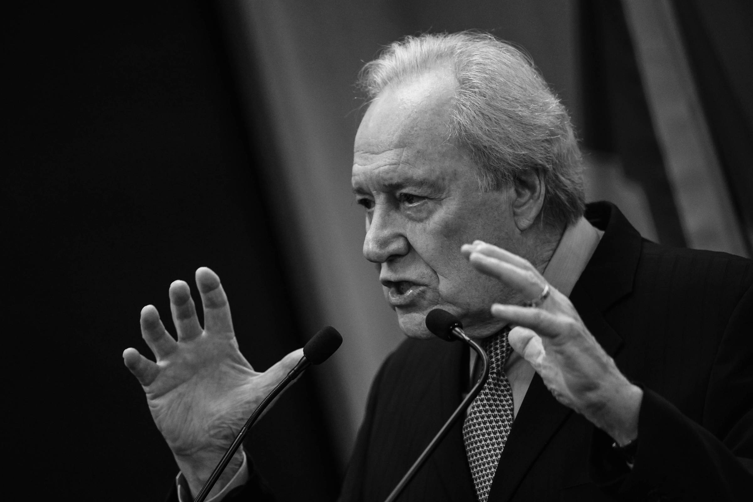 Um homem de cabelos grisalhos e com um terno escuro está falando em um microfone. Ele gesticula com as mãos, expressando-se de forma enfática. Ao fundo, há uma bandeira do Brasil visível, com as cores verde e amarelo.