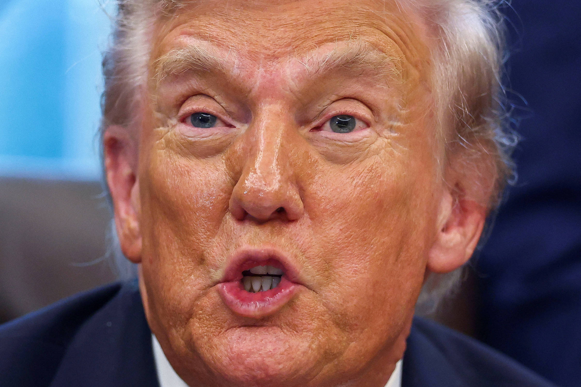Retrato próximo de homem branco com cabelo loiro claro e pele bronzeada, com expressão facial enquanto fala. Fundo desfocado em tons azul e escuro.