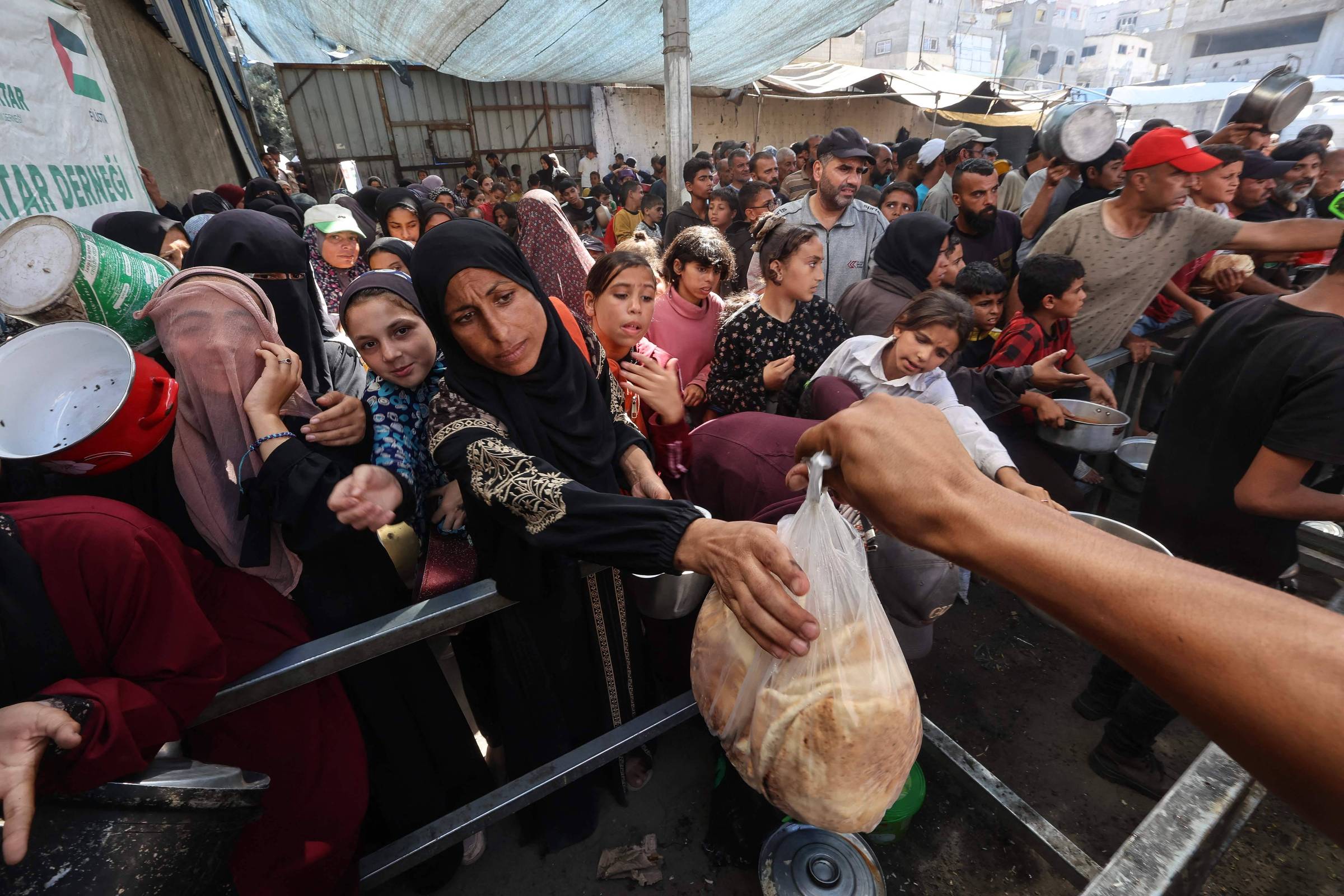 Grande grupo de pessoas, incluindo mulheres e crianças, aguardam em fila sob cobertura improvisada para receber alimentos. Uma mão entrega um saco plástico com pão para uma mulher na frente da fila.