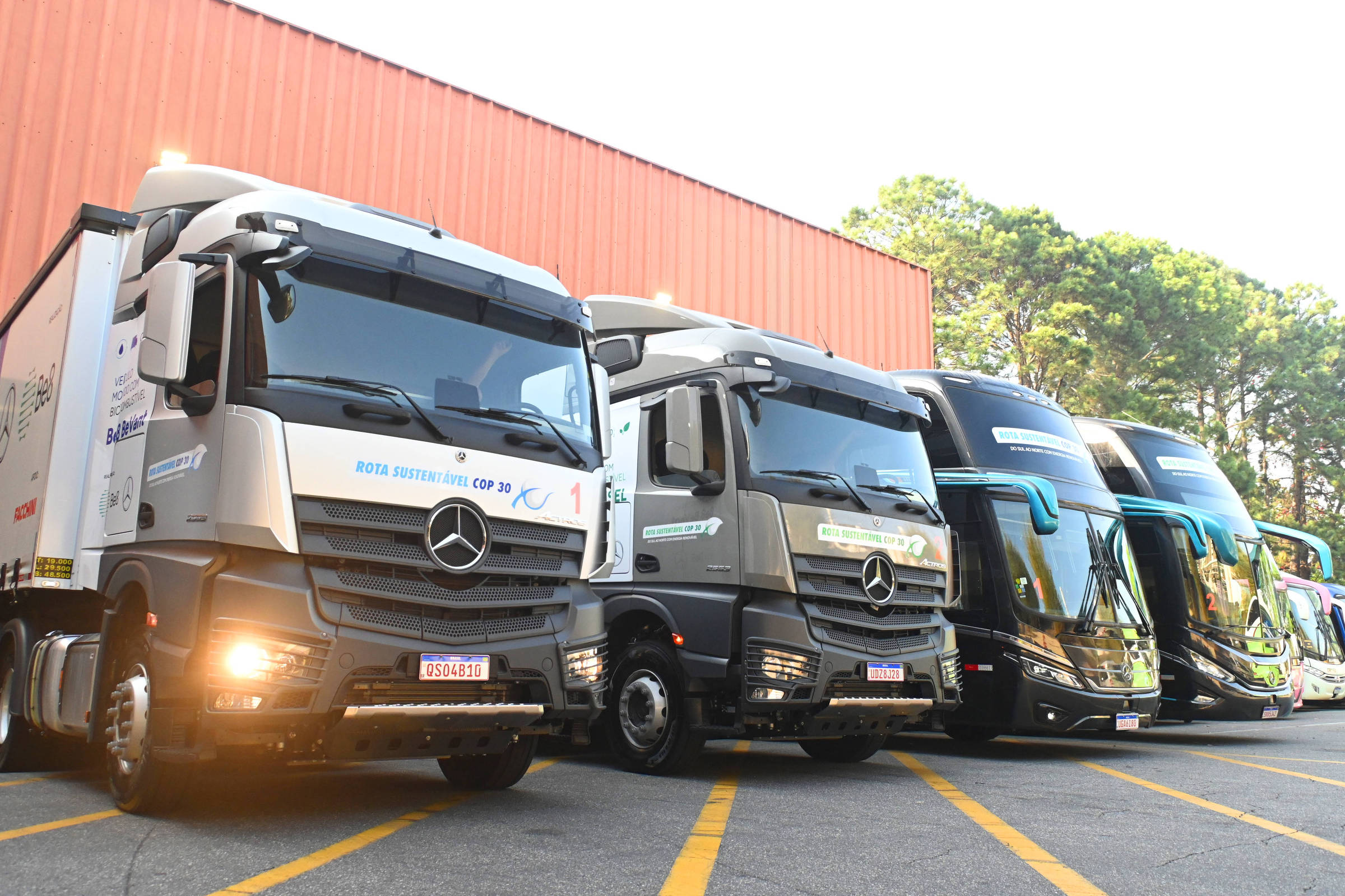 Quatro caminhões Mercedes-Benz alinhados em estacionamento externo, com cabine branca, cinza e duas pretas. Fundo com parede laranja e árvores ao fundo.