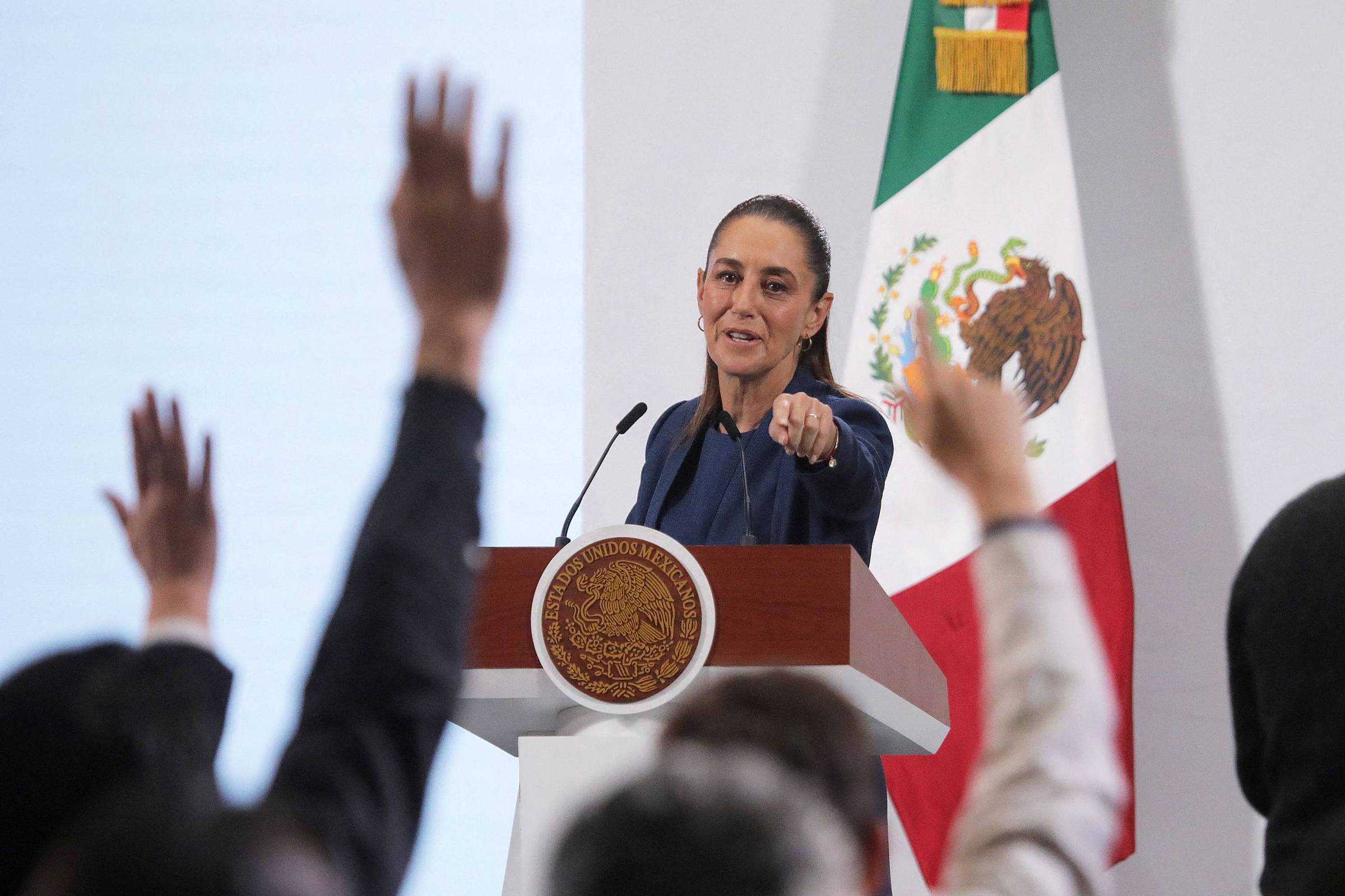 Prefeita em púlpito com brasão oficial fala para público que levanta as mãos. Bandeira do México está ao fundo, e câmera registra o evento.