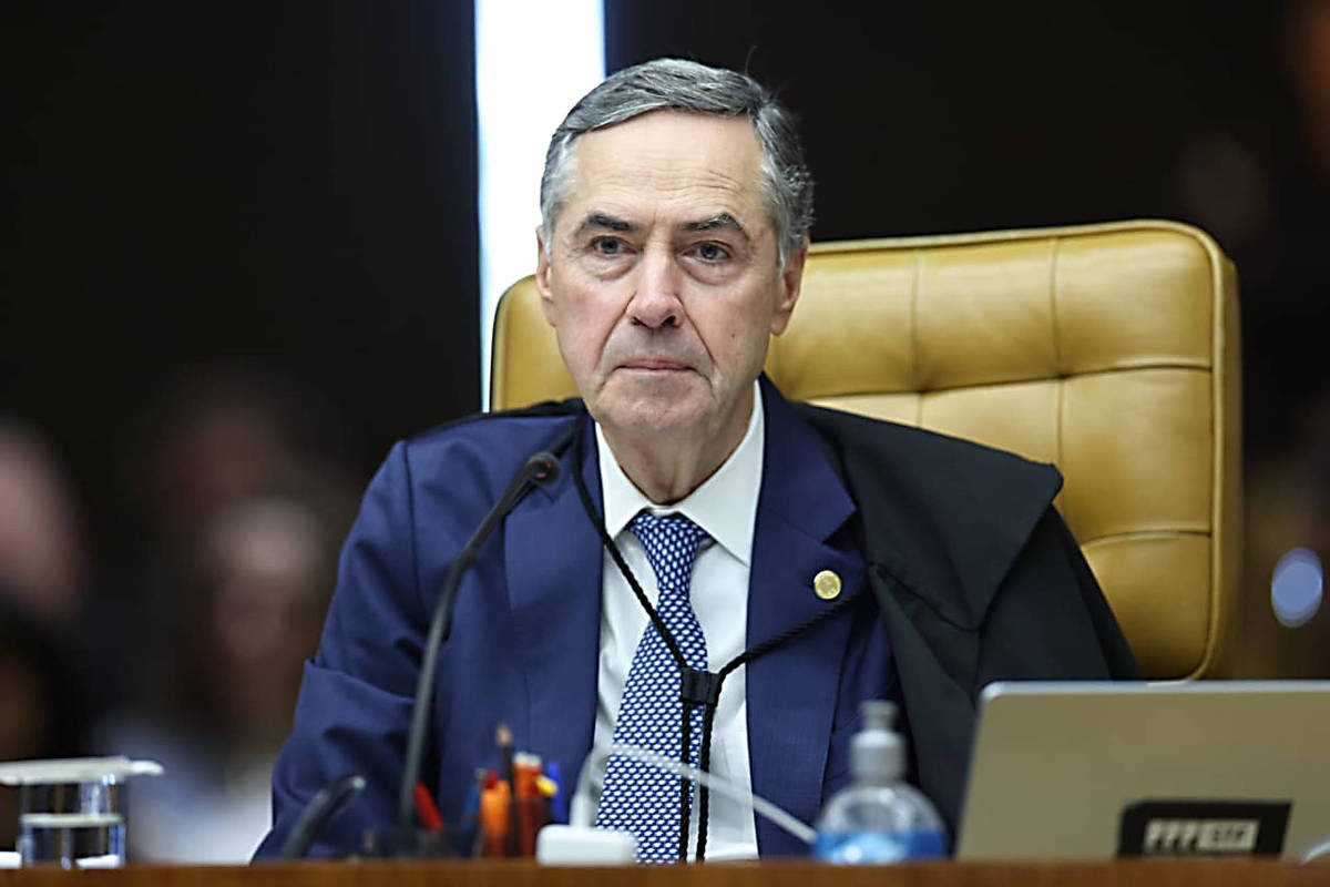 Ministro do Supremo Tribunal Federal sentado em cadeira de couro bege, vestindo terno azul escuro, camisa branca e gravata azul com padrão. Microfone à frente e laptop aberto ao lado direito. Fundo escuro com luz vertical branca ao centro.