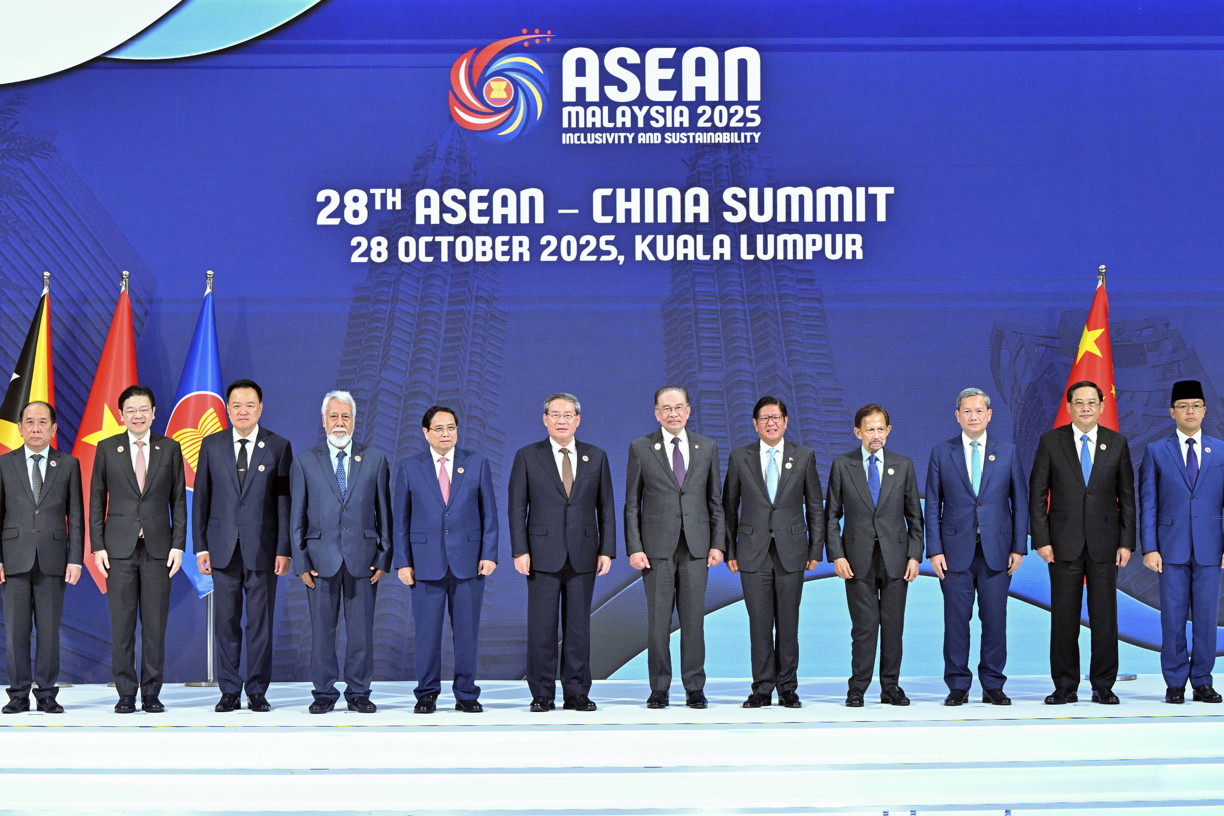 Doze líderes posam alinhados no palco da 28ª Cúpula ASEAN-China em Kuala Lumpur, em 28 de outubro de 2025. Ao fundo, painel azul com o logo do evento e bandeiras nacionais dos países participantes à esquerda e direita.