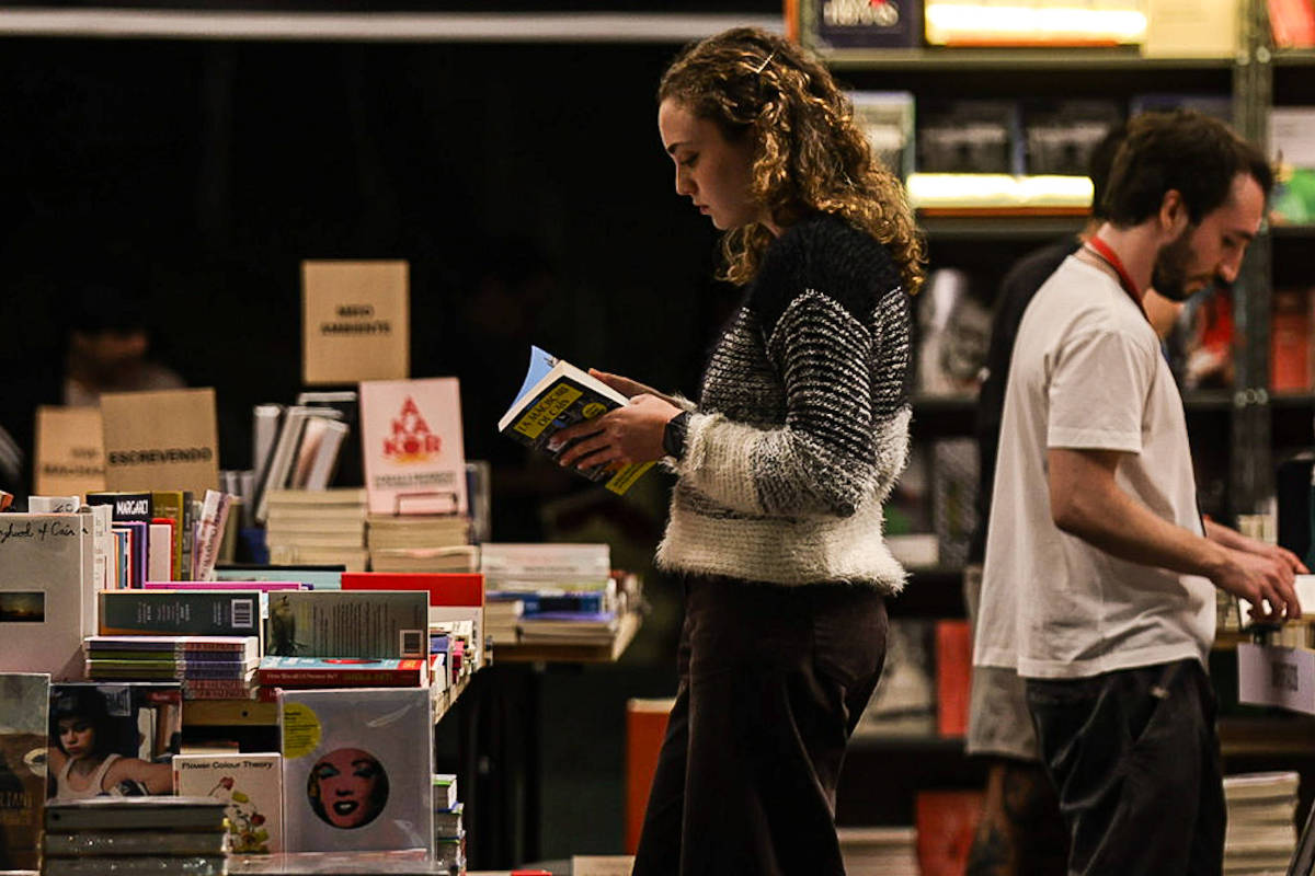 Mulher com suéter listrado lê livro em estande de feira de livros. Homem ao fundo examina outros livros em prateleira.