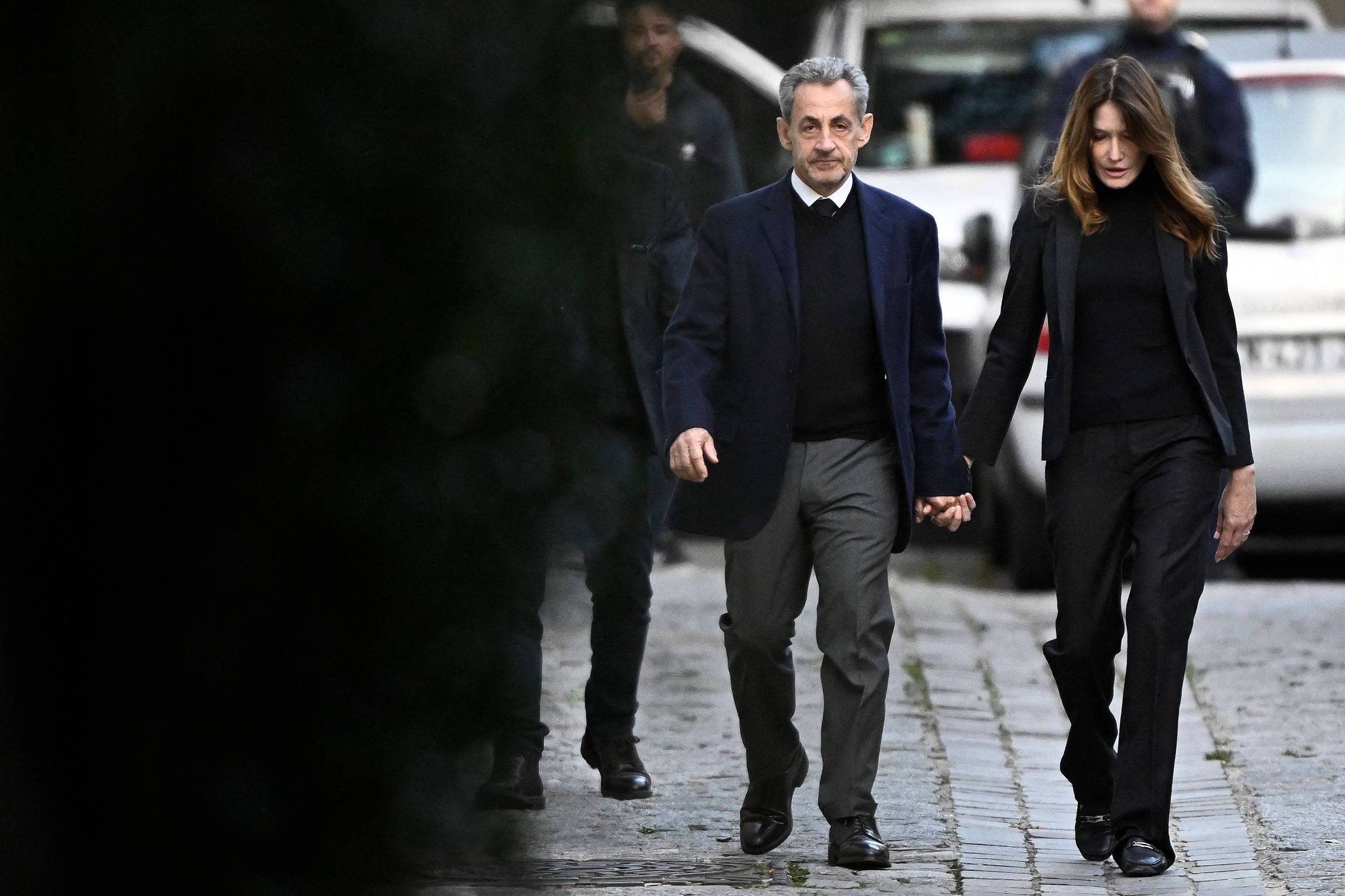 Homem e mulher vestidos com roupas escuras caminham de mãos dadas por uma rua de paralelepípedos. Ao fundo, duas pessoas e um veículo branco estão visíveis.