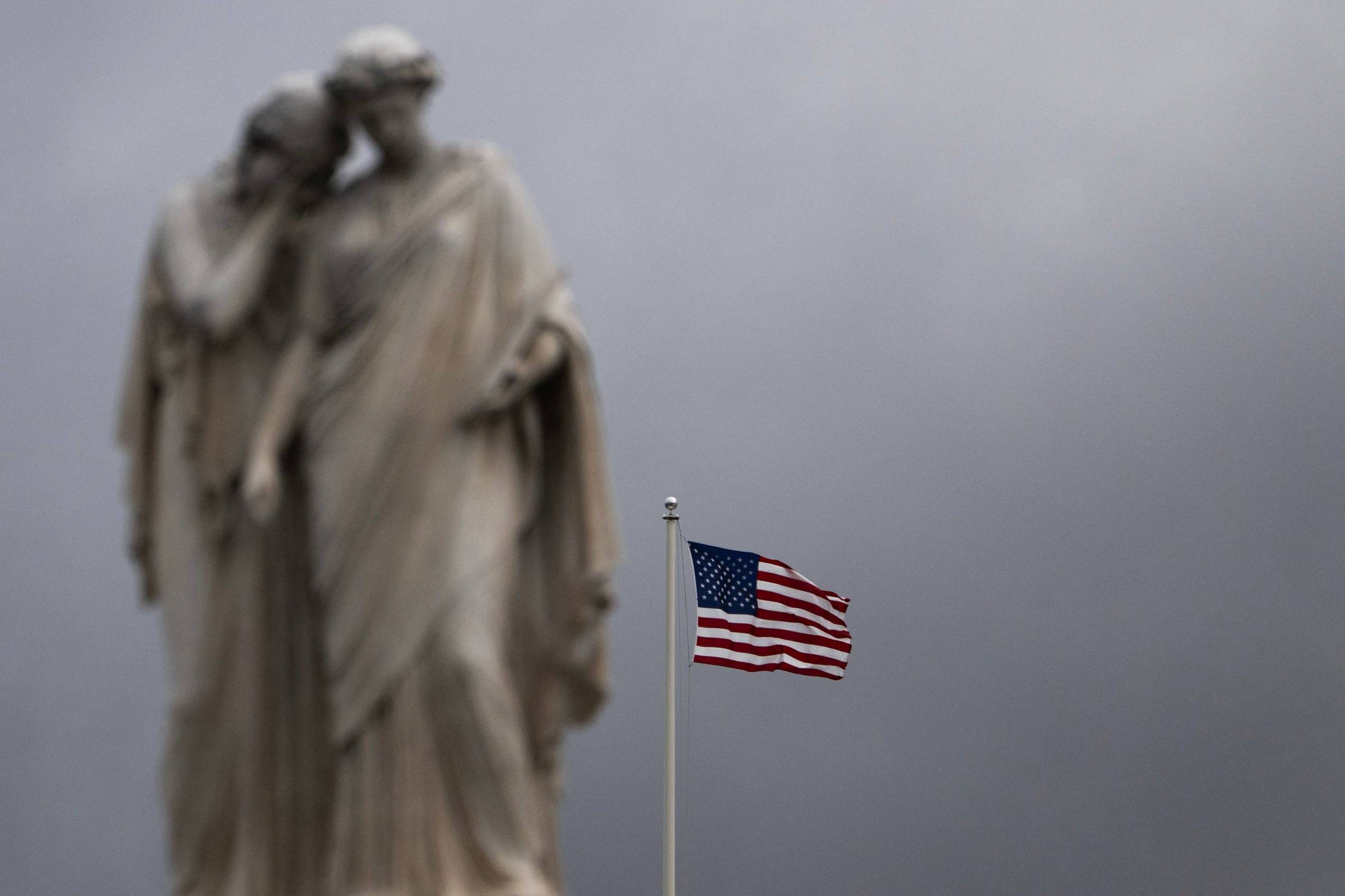 Bandeira dos Estados Unidos tremula em mastro contra céu nublado. Ao fundo, estátua de duas figuras humanas em pedra, desfocada.