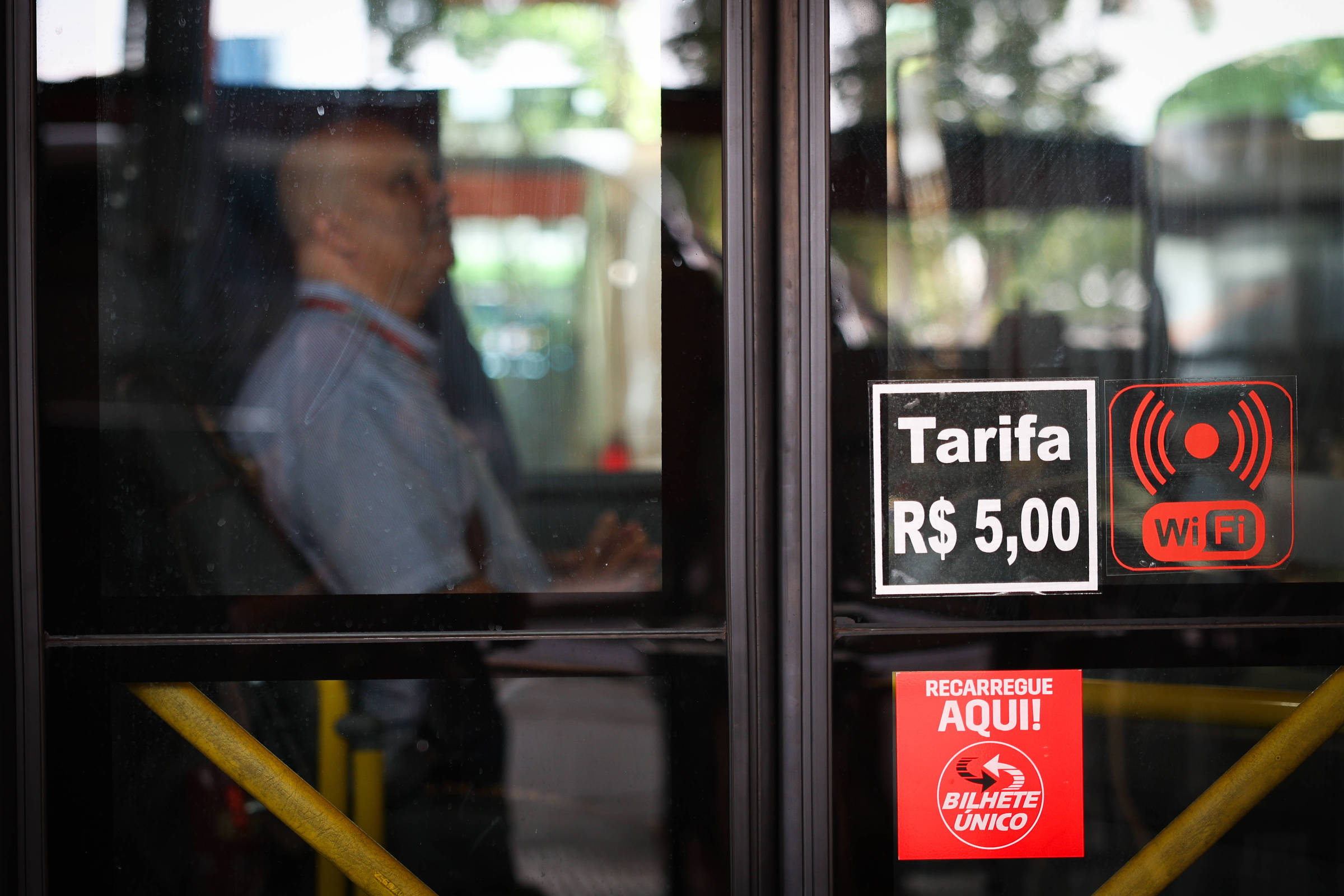Passageiro sentado dentro de ônibus visto através da porta de vidro. Na porta, adesivos indicam tarifa de R$ 5,00, disponibilidade de Wi-Fi e ponto para recarga do Bilhete Único.