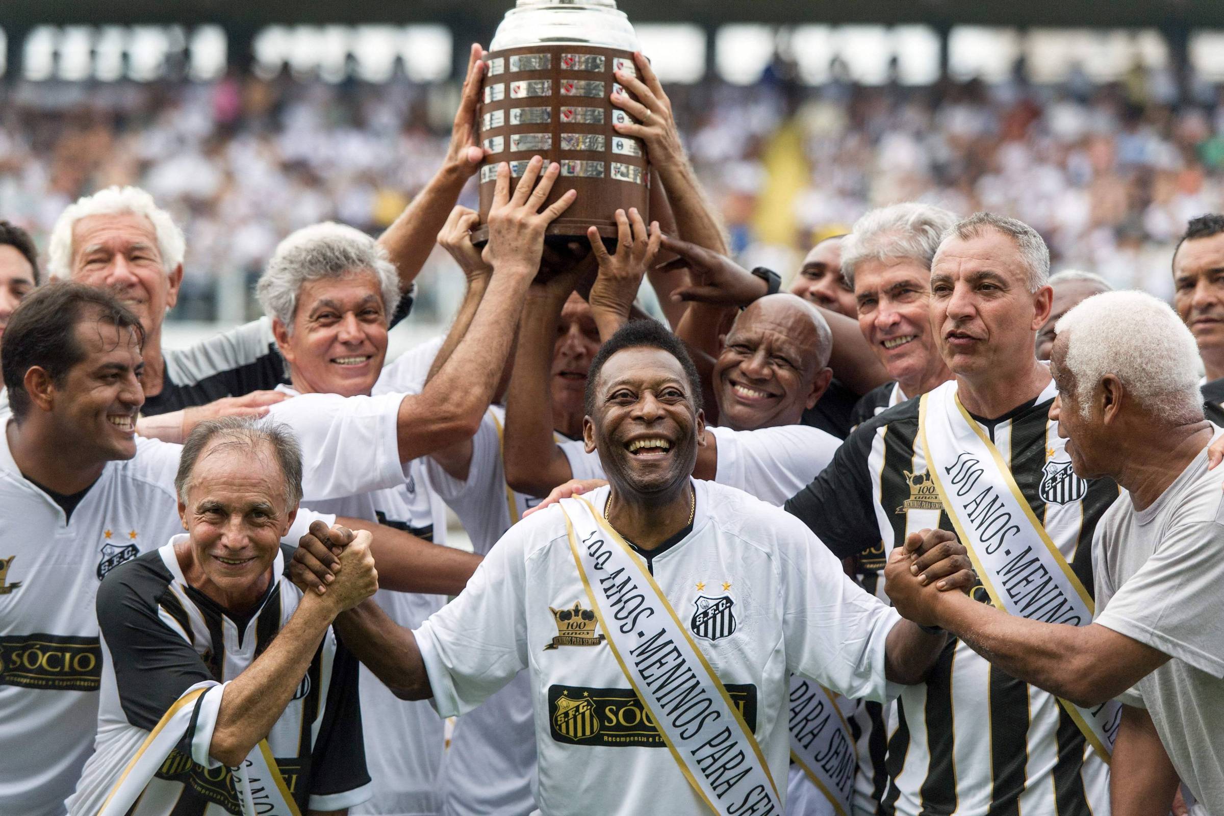 Grupo de ex-jogadores do Santos ergue a taça da Copa Libertadores em campo, com Pelé ao centro sorrindo e usando faixa comemorativa. Público desfocado ao fundo.